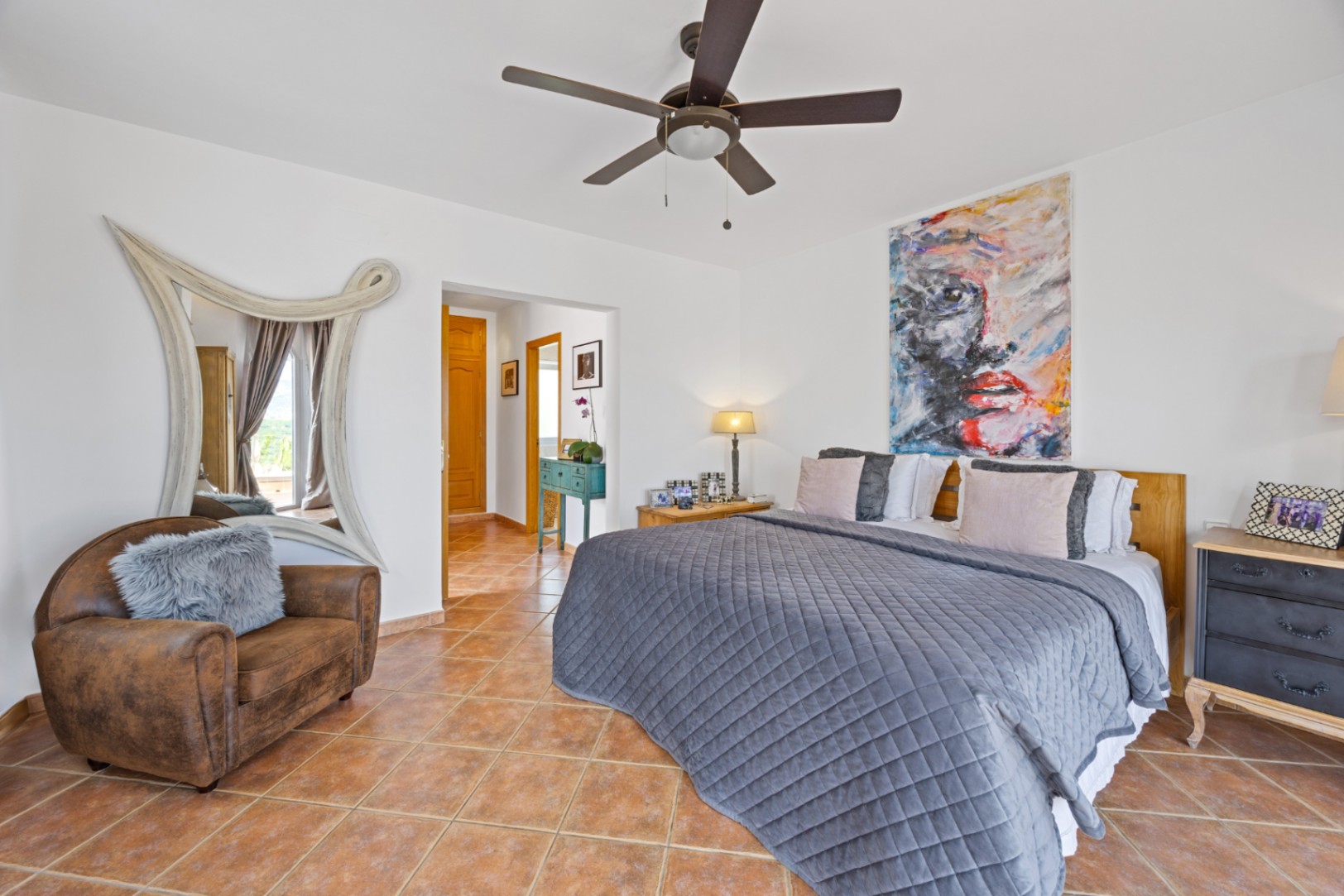 Dormitorio con suelos de terracota, un gran retrato abstracto y un espejo de marco blanco ornamentado. Incluye ventilador de techo y un sillón de cuero con cojín de pelo.
