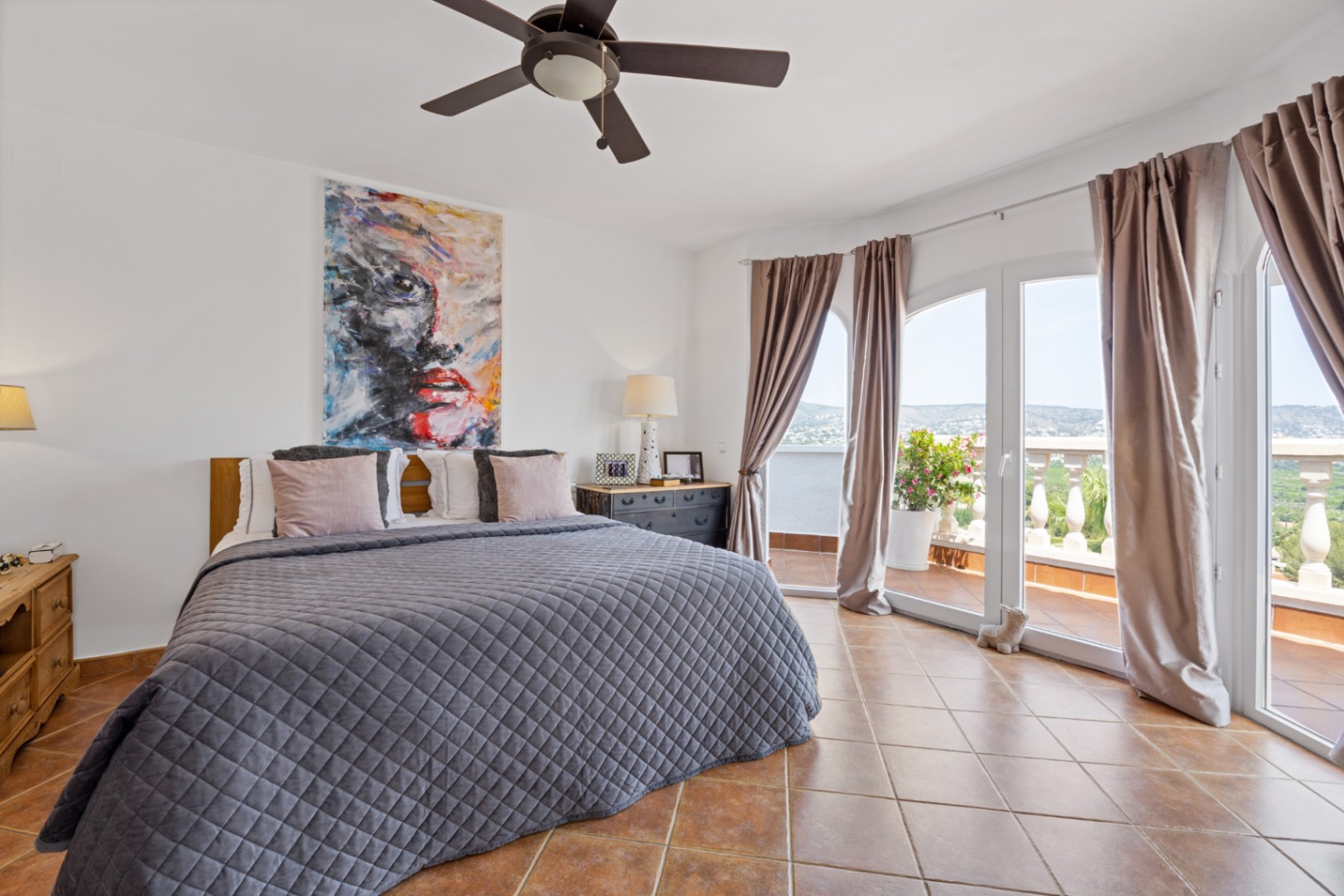 Dormitorio con suelos de terracota, ventilador de techo y grandes ventanales de arco que comunican con una terraza con vistas panorámicas.