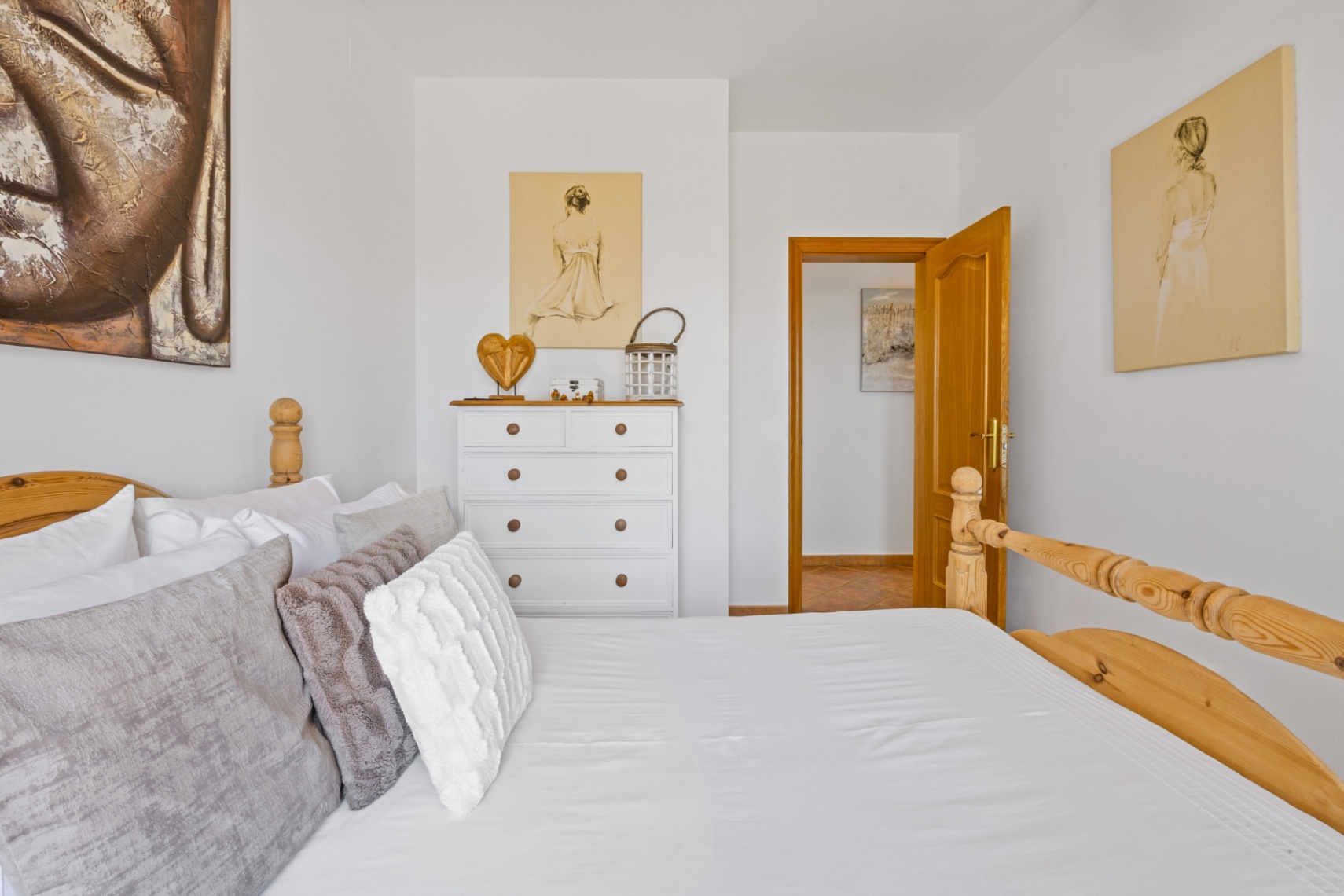 Dormitorio luminoso con cama de madera tallada, cómoda blanca con detalles naturales y paredes decoradas con arte figurativo y un lienzo de Buda.