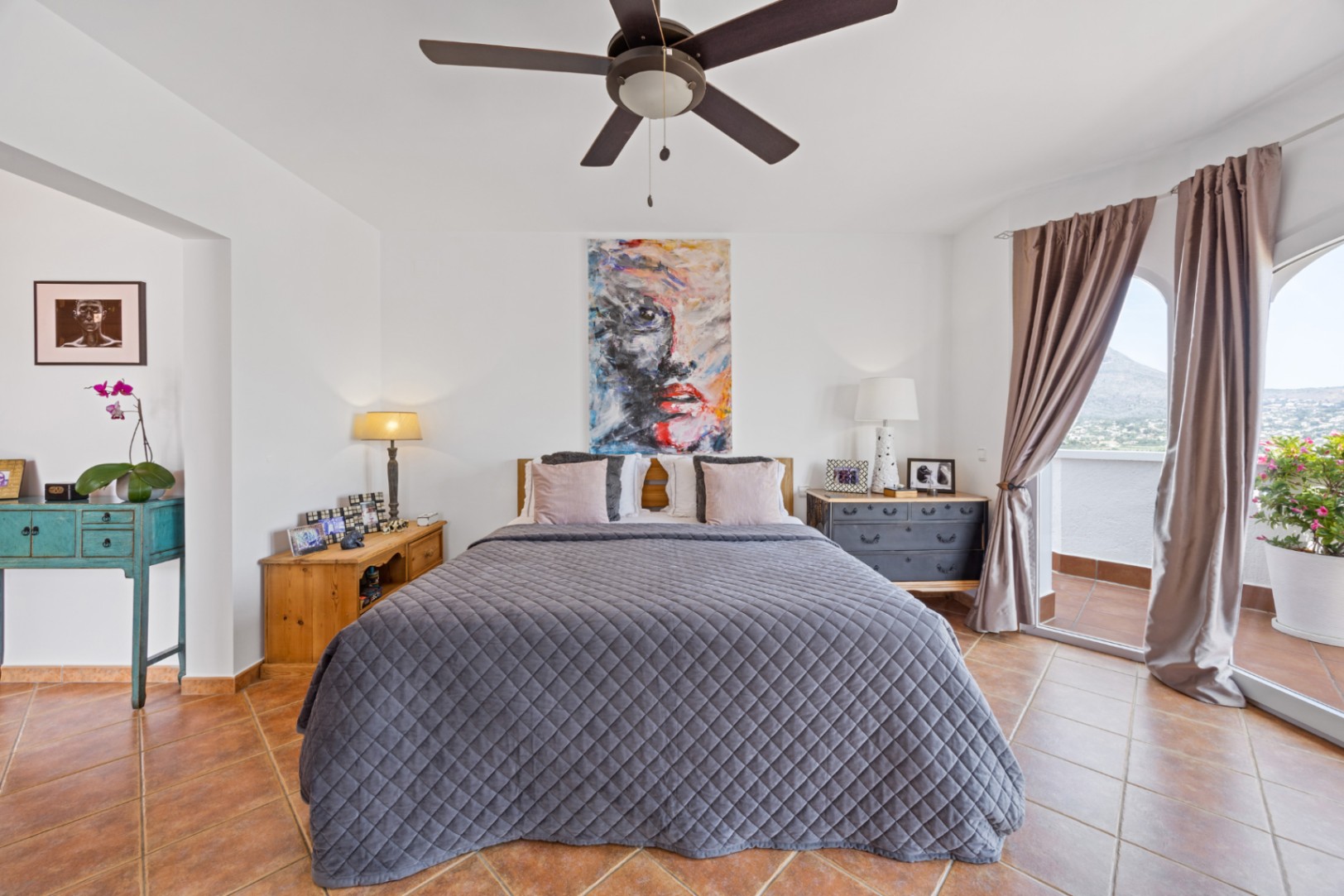 Dormitorio luminoso con suelos de terracota, ventilador de techo y puertas correderas de cristal con acceso a un balcón privado y vistas a la montaña.