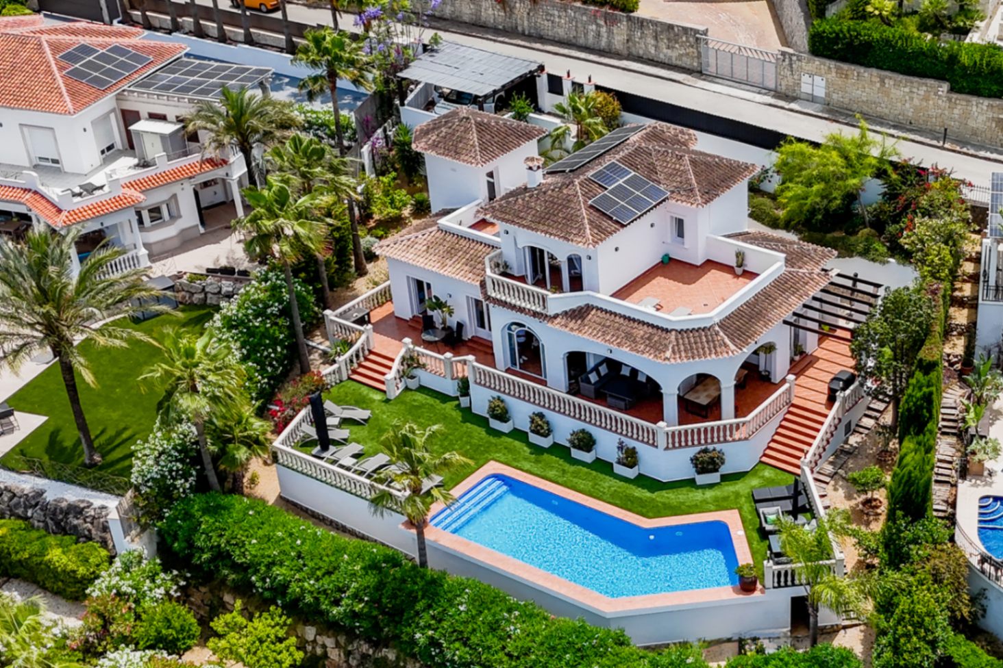 Perspectiva aérea de una villa mediterránea con piscina geométrica, paneles solares y terrazas de terracota en varios niveles con balaustradas blancas.