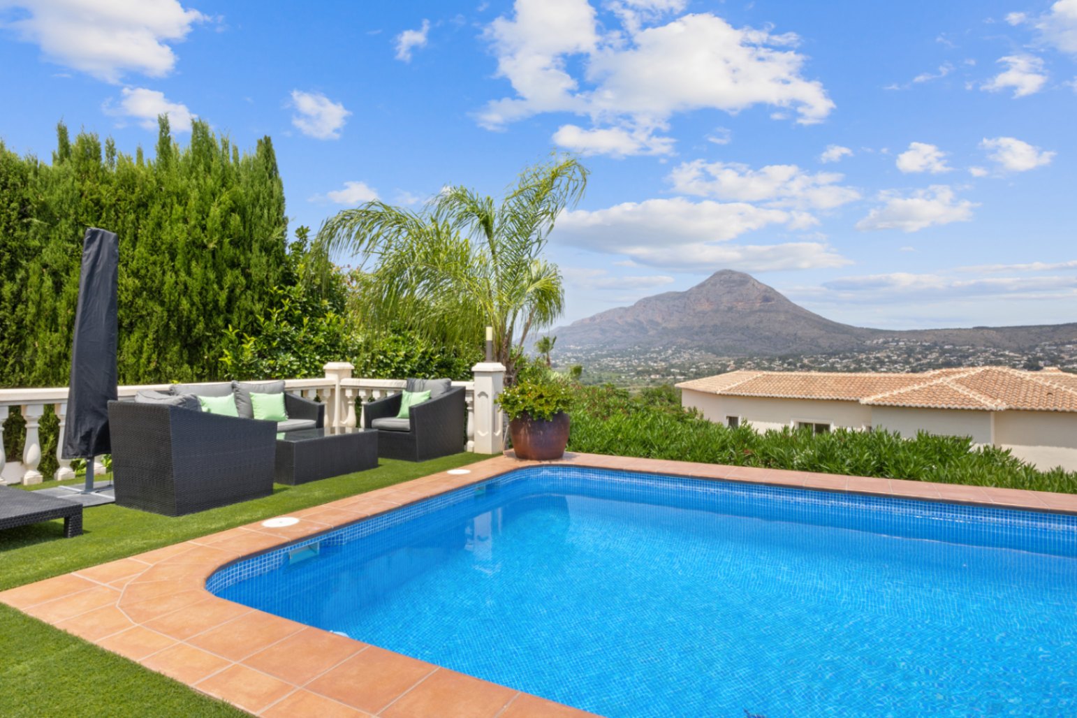 Piscina exterior con borde de terracota, zona de estar con césped artificial y muebles de mimbre, ofreciendo vistas despejadas a las montañas.