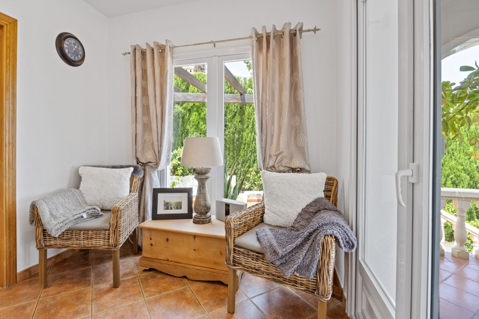 Rincón de descanso con dos sillones de mimbre, baúl de madera rústico y suelos de terracota. El ventanal ofrece vistas directas al jardín y a la pérgola exterior.