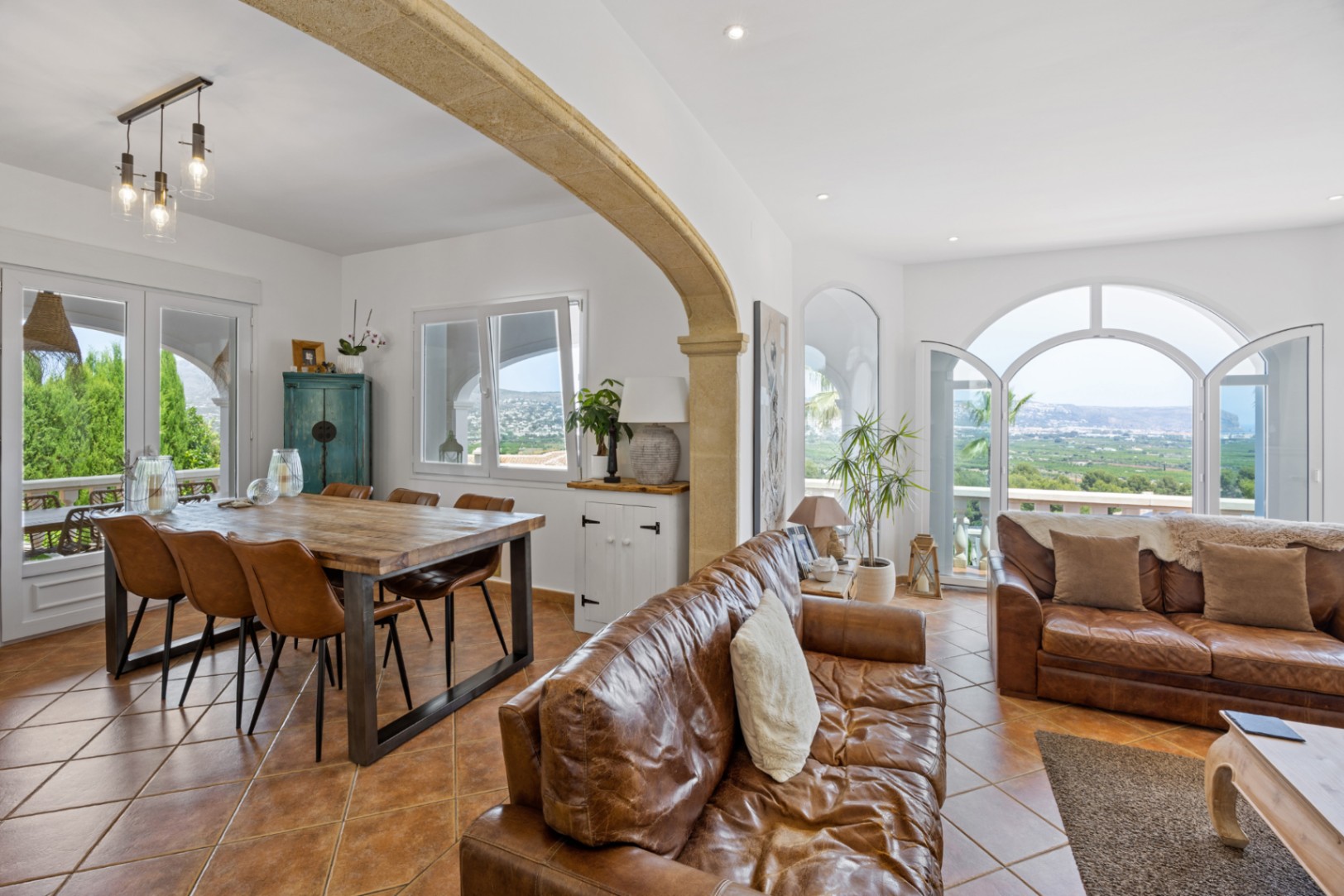 Salón-comedor diáfano con arco de piedra natural, sofás de cuero y mesa de madera. Grandes ventanales en arco ofrecen vistas despejadas al valle.