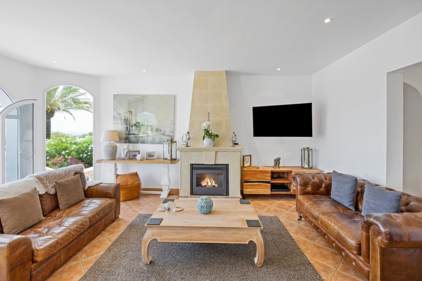 Salón con chimenea de piedra, sofás de cuero marrón y suelos de terracota. Los ventanales en arco conectan el interior con un jardín mediterráneo exterior.
