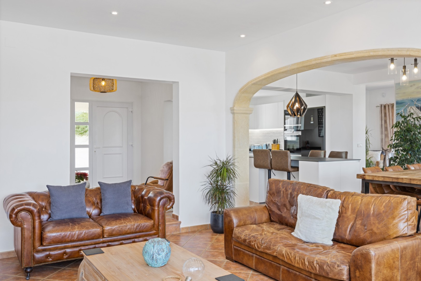 Salón de planta abierta con sofás de cuero, suelos de terracota y un arco de piedra natural que conecta con una cocina moderna y zona de comedor.