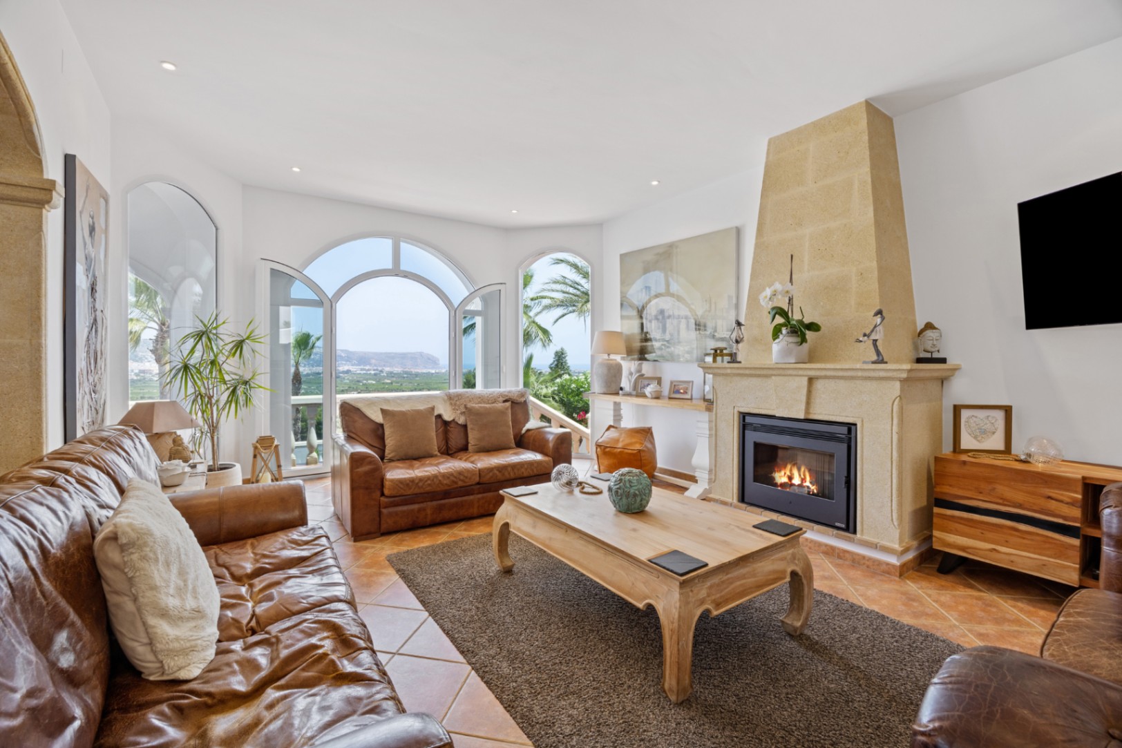 Salón elegante con chimenea de piedra, sofás de cuero y ventanales en arco que conectan con una terraza con vistas al mar y la montaña.