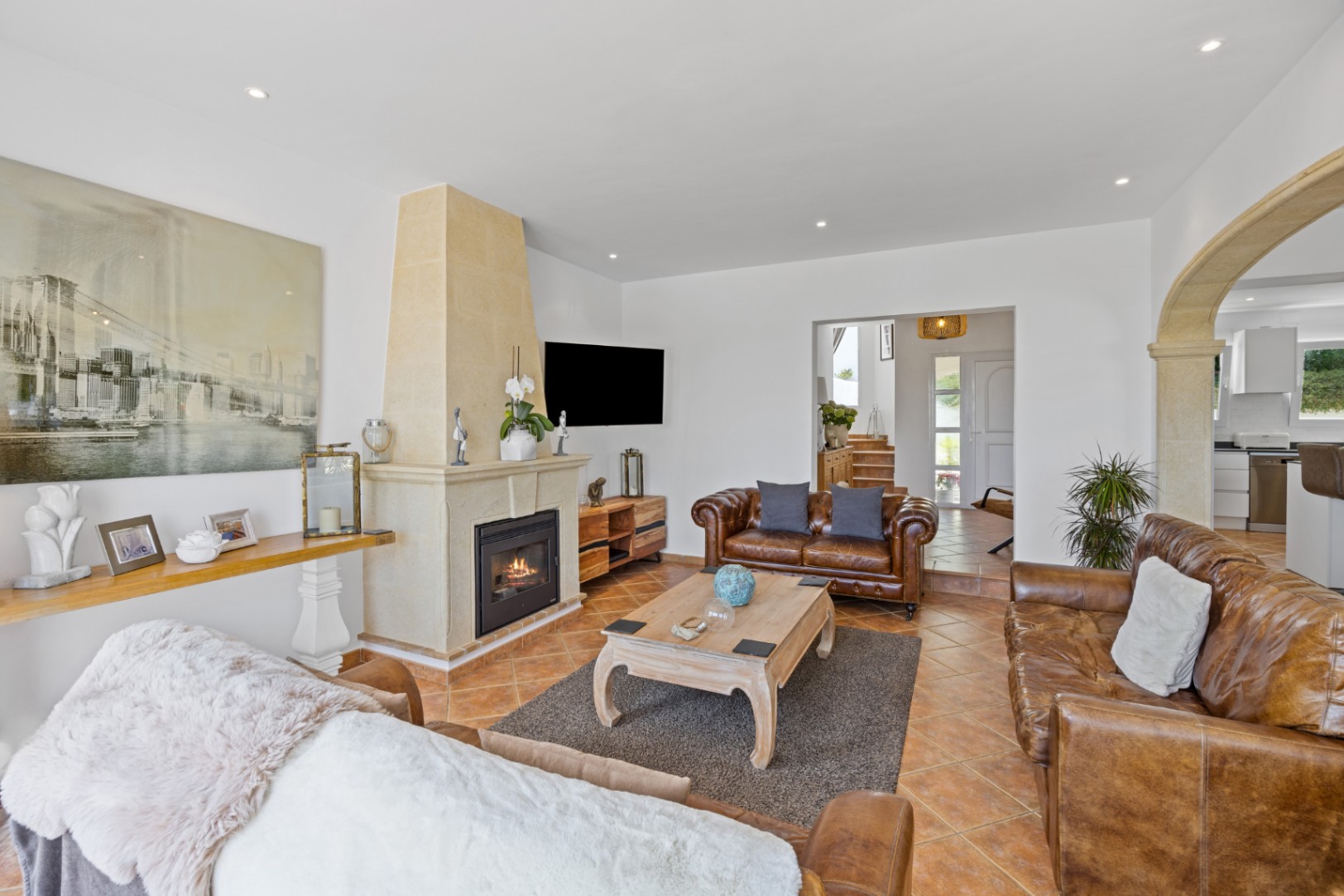 Salón espacioso con sofás de cuero marrón, chimenea de piedra natural y suelos de terracota. Un arco de piedra comunica la estancia con la cocina y el recibidor.