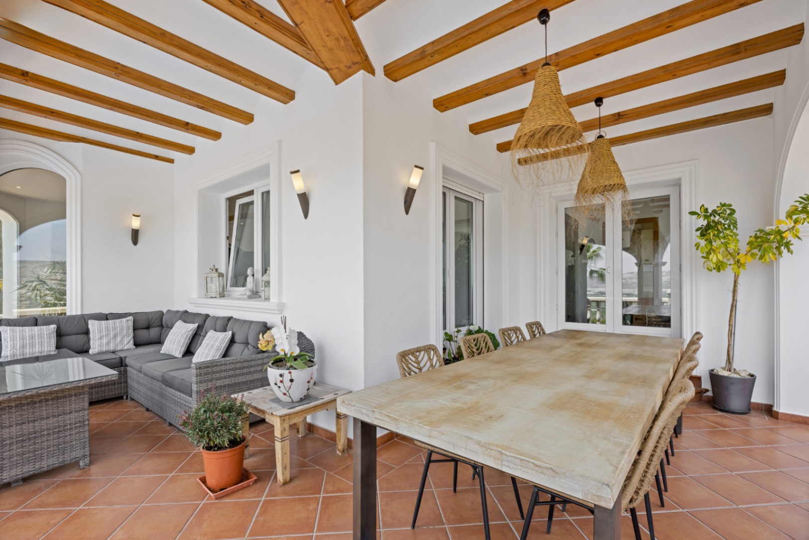 Terraza cubierta con vigas de madera, suelo de terracota, mesa de comedor de madera con sillas trenzadas y zona de estar con sofá de mimbre gris.