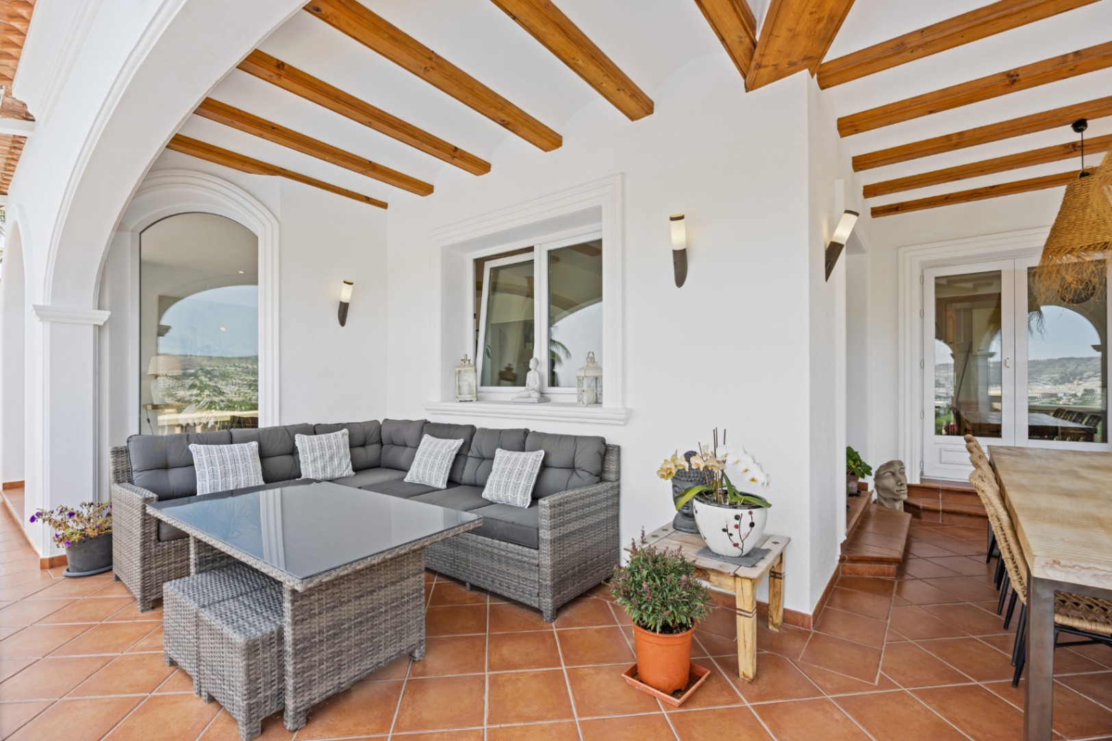 Terraza cubierta con vigas de madera, suelo de terracota y mobiliario de mimbre gris. Incluye apliques de pared y vistas despejadas a través de los arcos.