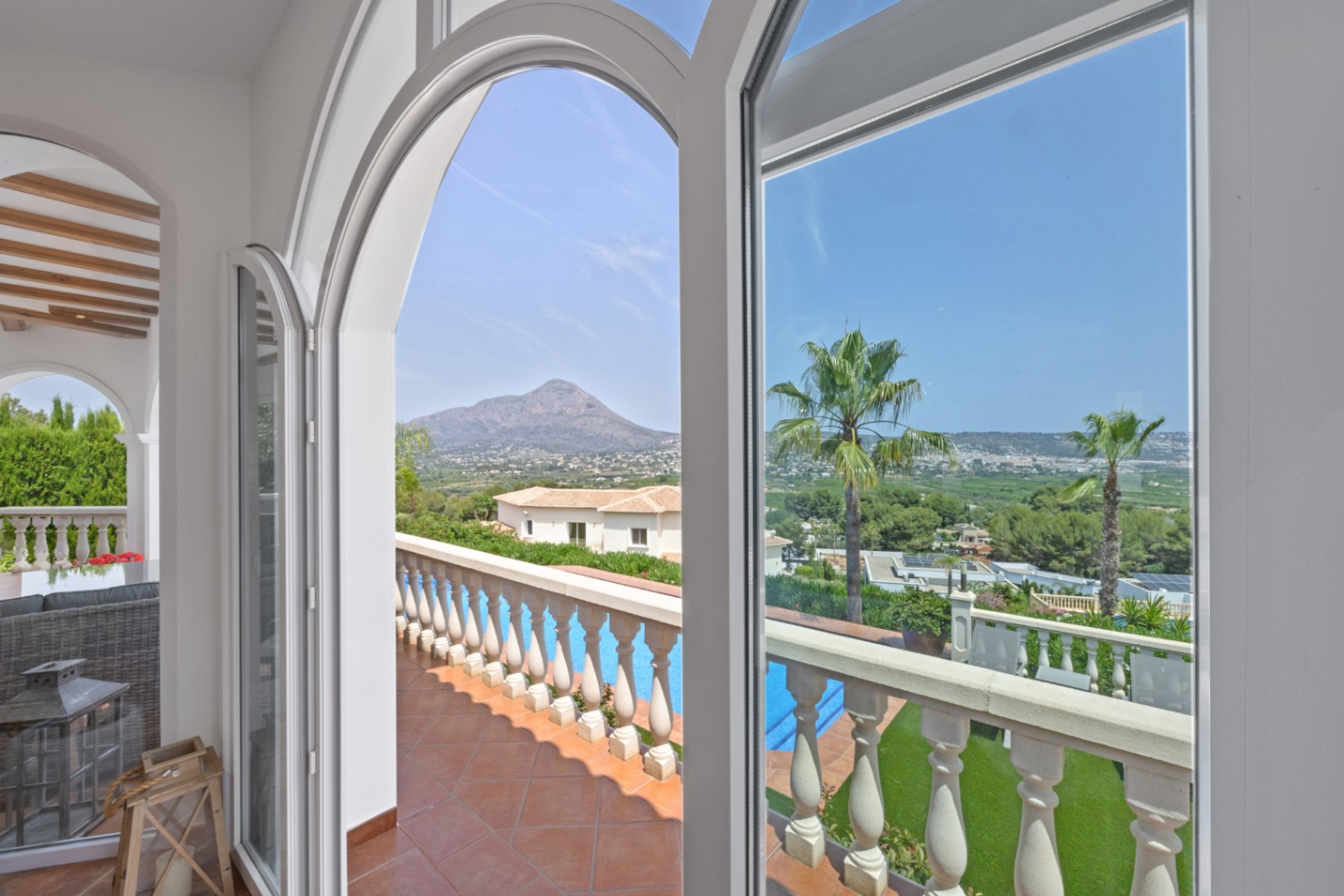Ventanales de arco blanco enmarcan vistas panorámicas a la montaña y al valle, con acceso a una terraza de terracota y balustrada frente a la piscina.