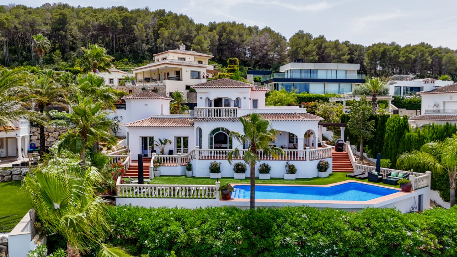 Villa mediterránea de fachada blanca y tejado de terracota, con terrazas multinivel, piscina privada y jardín con palmeras frente a una colina boscosa.