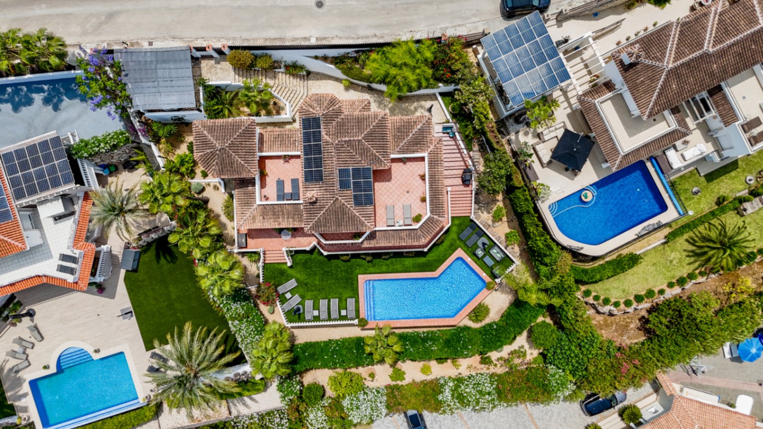 Vista aérea de una villa con tejado de terracota y paneles solares, piscina privada de forma geométrica y jardines paisajísticos con palmeras y terrazas.