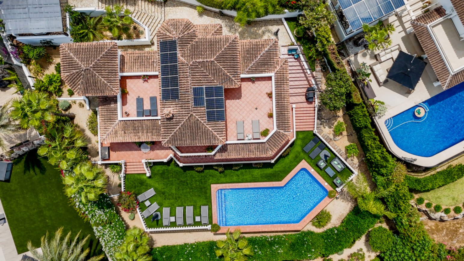 Vista aérea de una villa mediterránea con tejado de terracota, paneles solares, múltiples terrazas y una piscina de forma irregular rodeada de césped y vegetación.