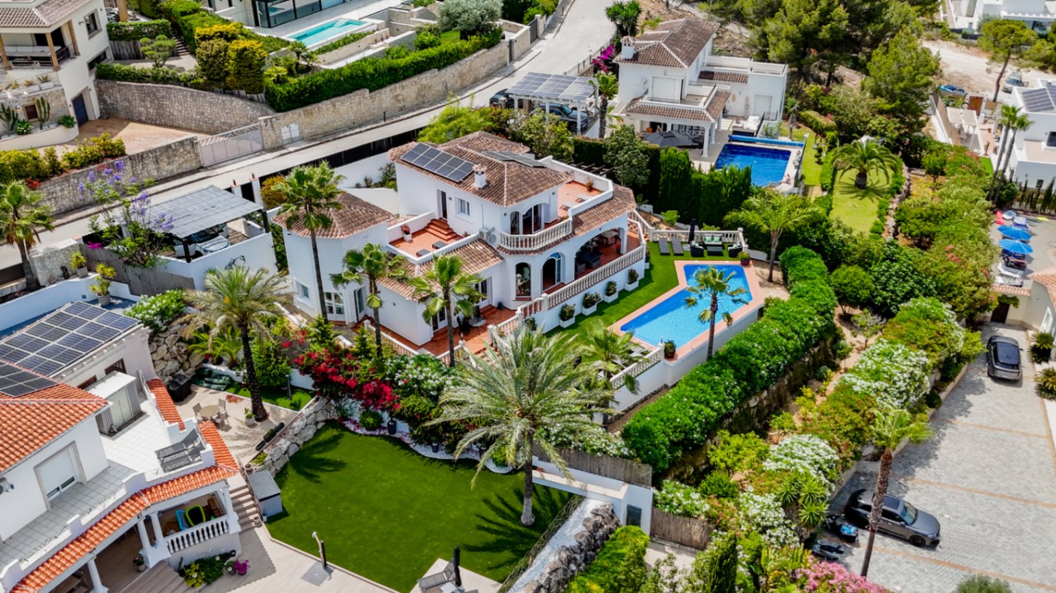 Vista aérea de una villa mediterránea con tejados de terracota, paneles solares, piscina privada de diseño geométrico y jardines con palmeras.