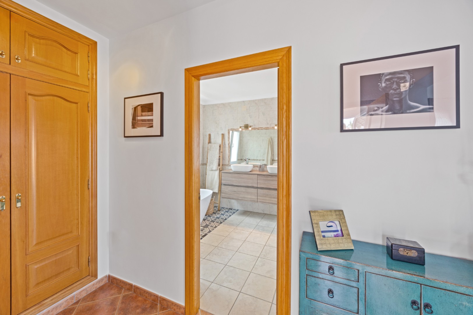 Vista desde el pasillo con armarios empotrados de madera y aparador azul, hacia un baño con lavabos de doble seno, mueble de madera y suelos cerámicos.