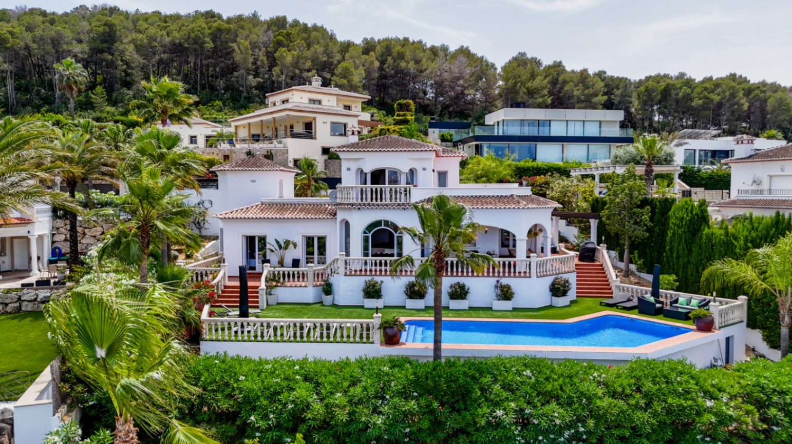 Vista elevada de una villa mediterránea blanca con tejados de terracota, ventanas de arco y piscina privada rodeada de palmeras y bosque de pinos.