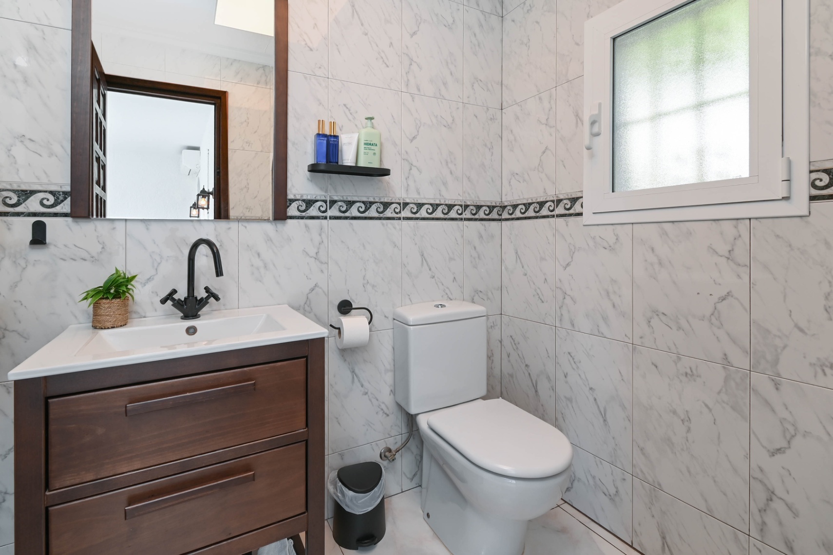 Baño con azulejos de efecto mármol y cenefa decorativa, mueble de madera oscura con lavabo blanco, grifería negra mate y ventana de cristal translúcido.