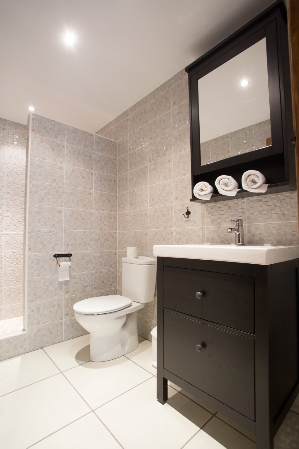 Baño moderno con paredes de mosaico en tonos neutros, mueble de lavabo en madera oscura y armario con espejo a juego que incluye estante para toallas.