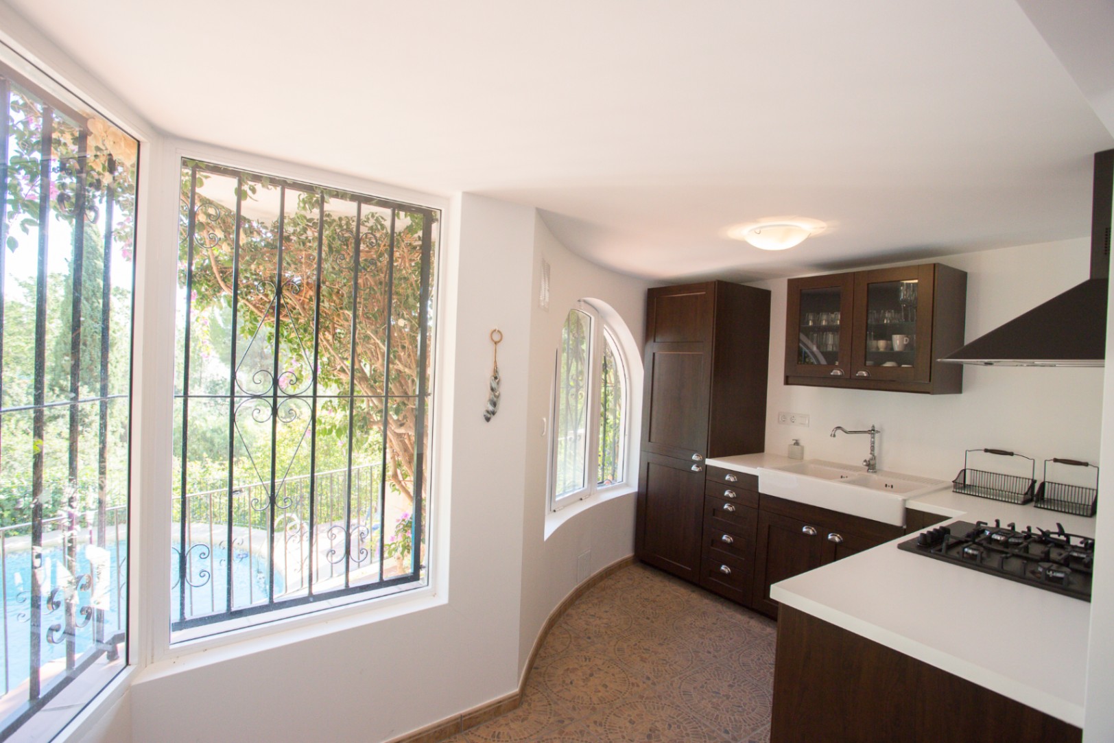 Cocina con armarios de madera oscura, encimeras blancas y fregadero de cerámica. Amplios ventanales con rejas decorativas ofrecen vistas al jardín y la piscina.