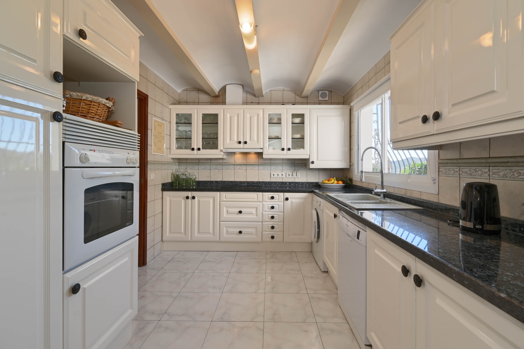 Cocina de estilo tradicional con mobiliario crema, encimeras de granito oscuro y techo abovedado con vigas de madera y electrodomésticos.