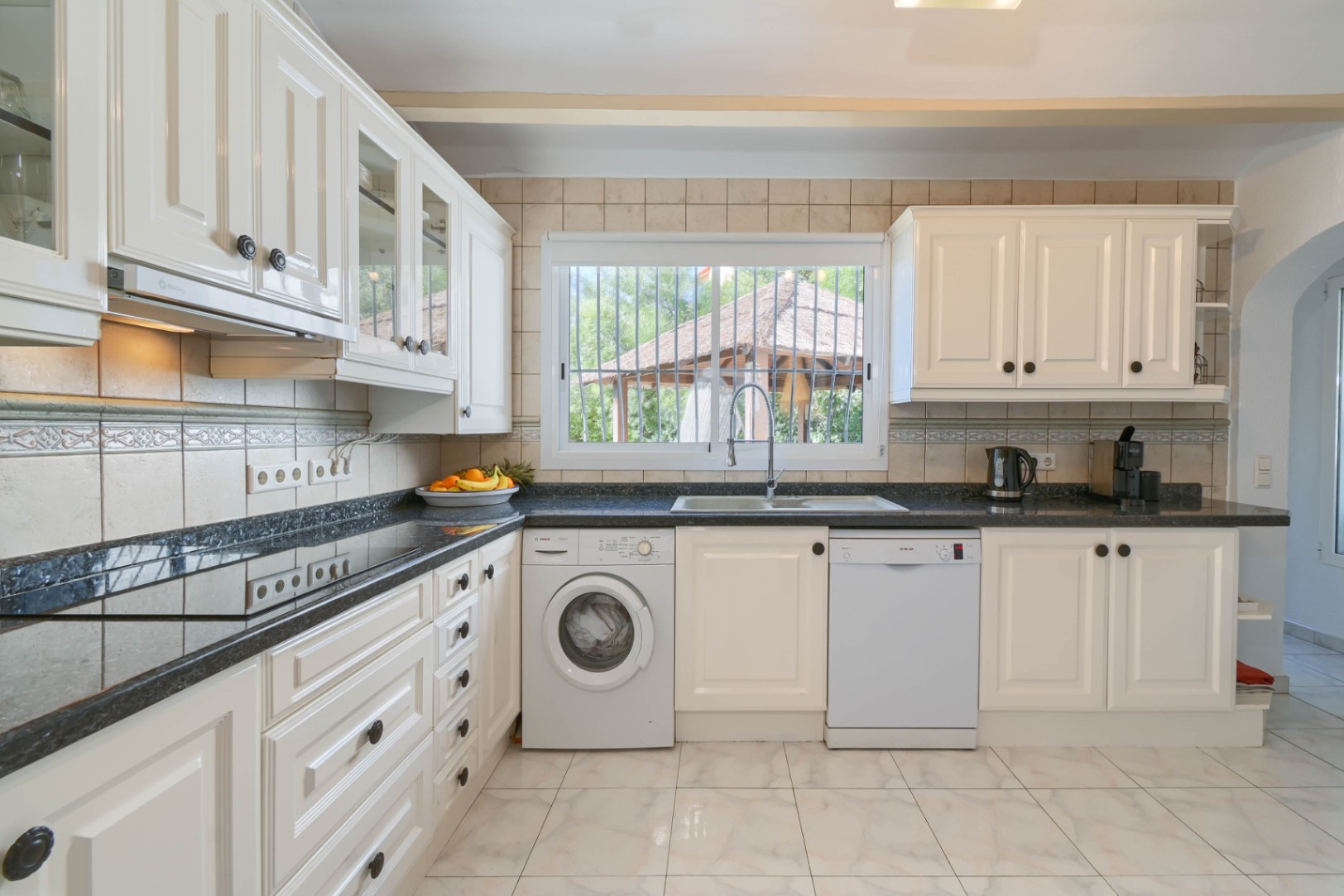 Cocina funcional con mobiliario blanco, encimera de granito oscuro y electrodomésticos integrados. Incluye zona de lavado y ventana con vistas al exterior.