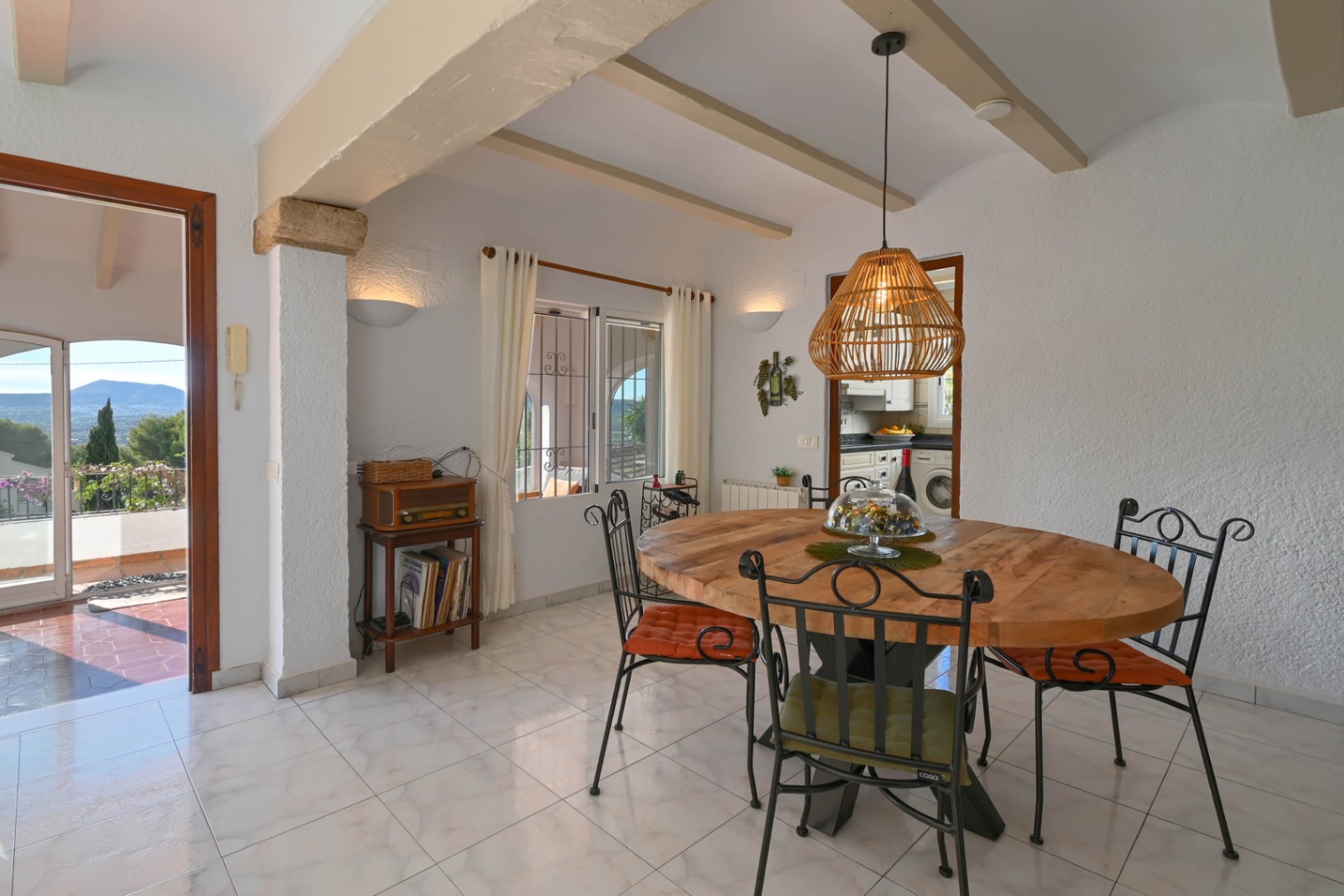 Comedor con mesa circular de madera, sillas de forja y lámpara de mimbre. Destacan las vigas vistas, suelos cerámicos y acceso directo a una terraza con vistas a la montaña.
