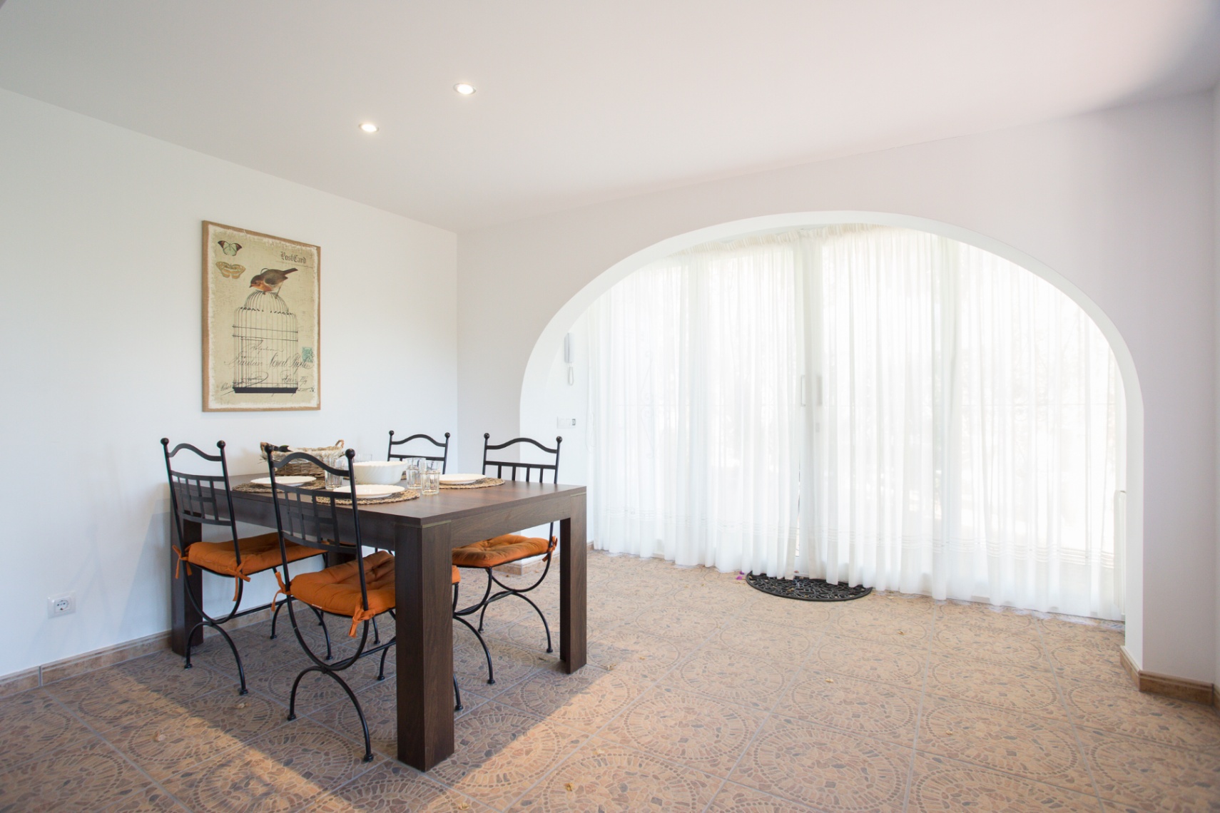 Comedor con mesa de madera oscura y sillas de forja con cojines naranjas. Destaca un gran arco con cortinas blancas y un suelo cerámico con motivos geométricos.