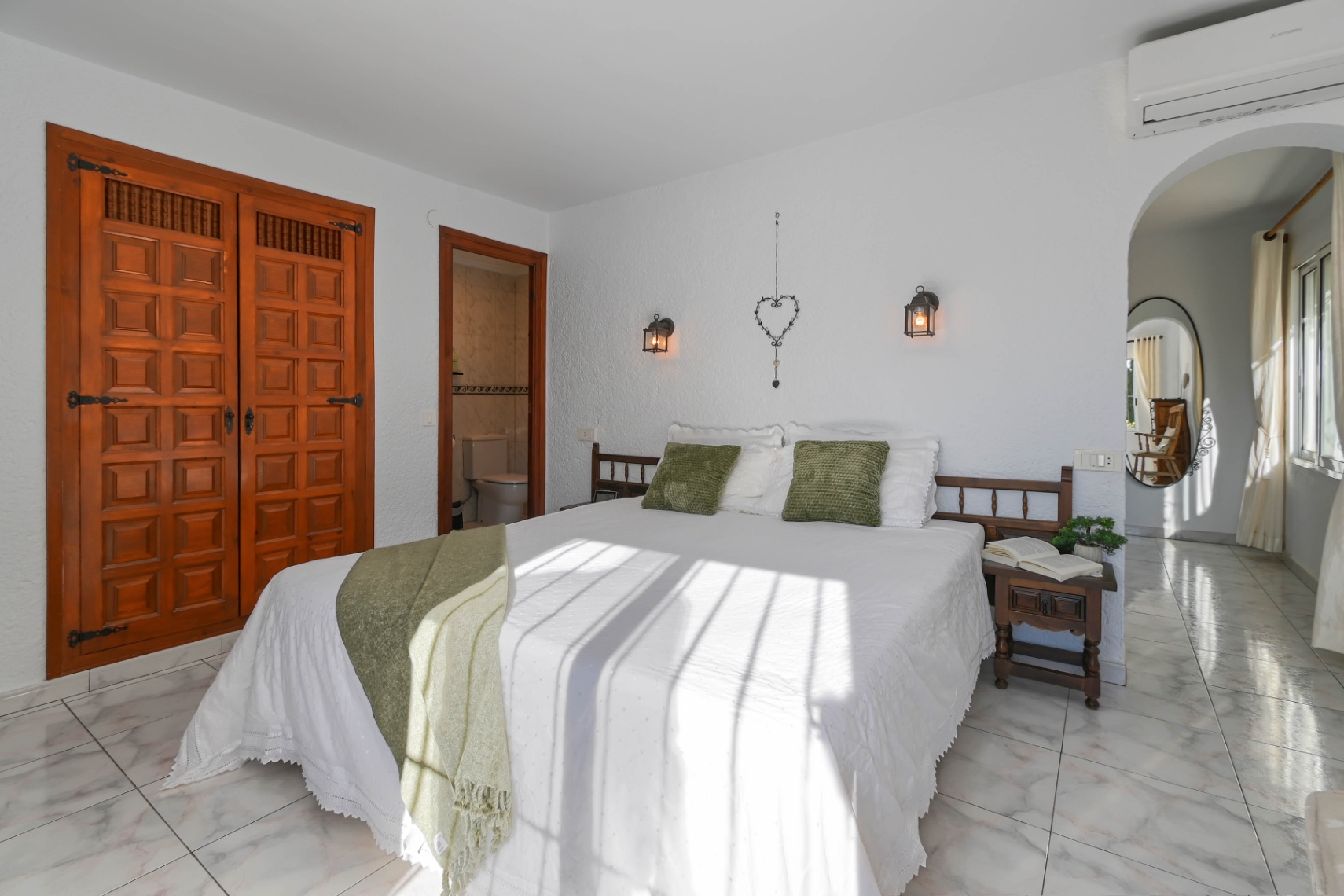 Dormitorio con armario empotrado de madera tallada, suelos de mármol claro y baño en suite. La luz natural ilumina la cama decorada con tonos verdes y apliques de estilo farol.