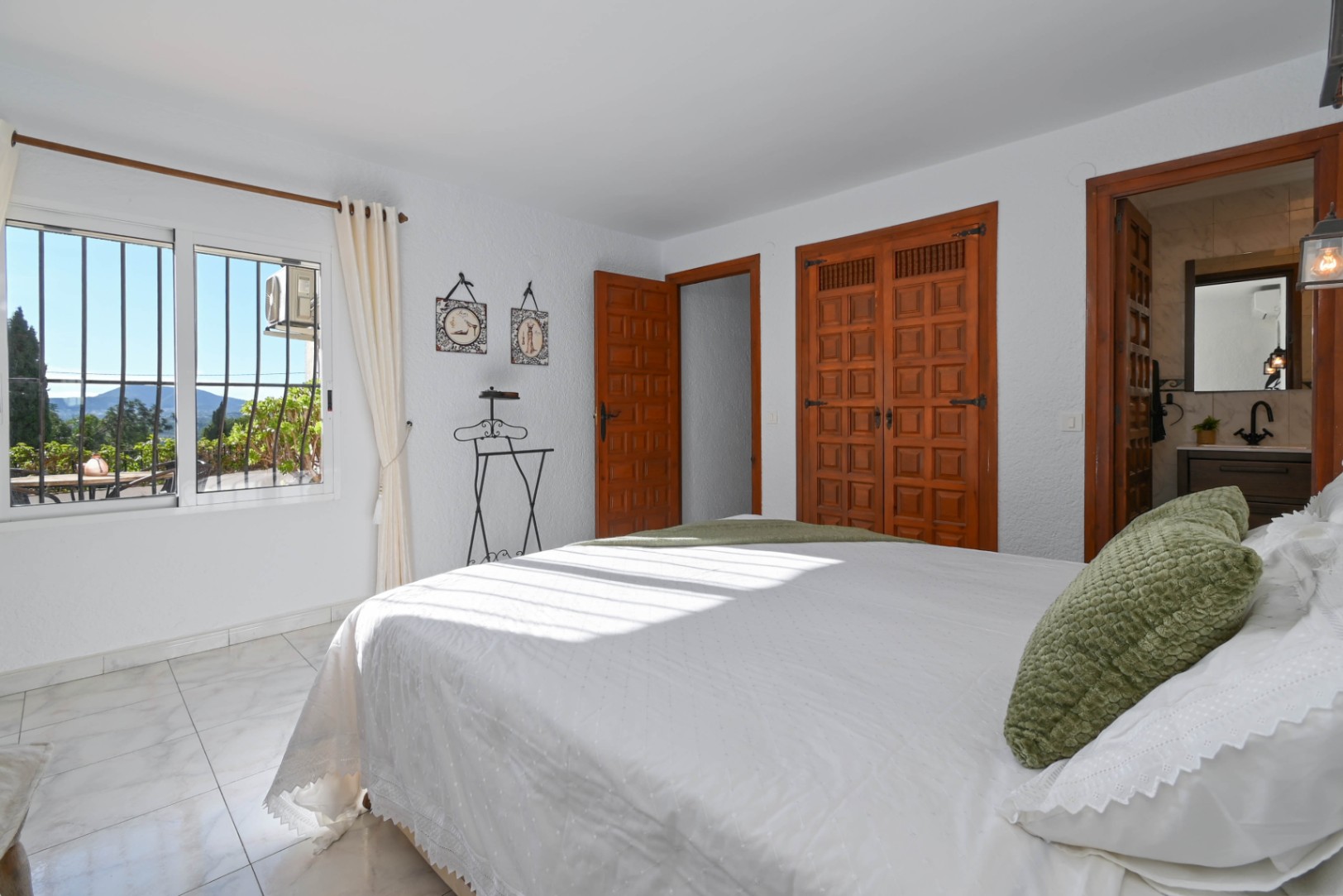 Dormitorio con armarios empotrados de madera tradicional, suelos de baldosa efecto mármol y baño en suite. El ventanal ofrece vistas despejadas al entorno natural.