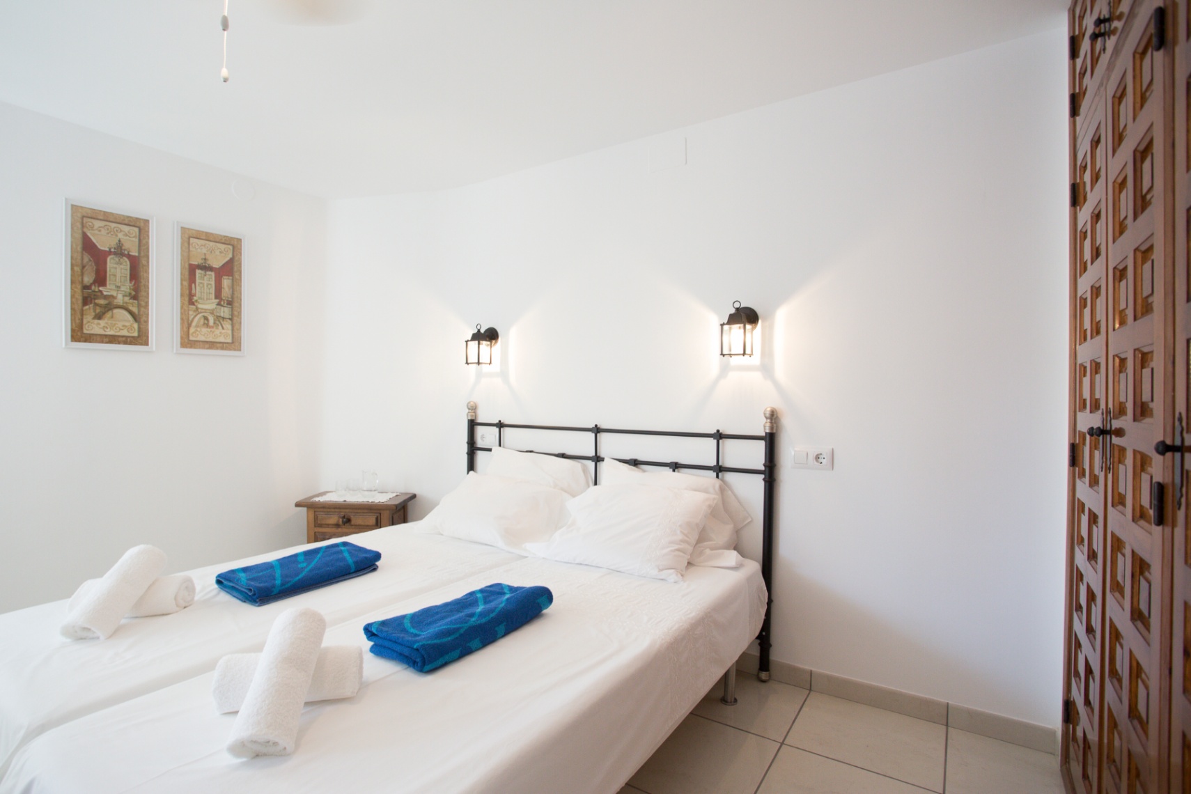Dormitorio con cabecero de forja negra, camas individuales con ropa blanca, apliques de pared tipo farol y un armario de madera tallada.