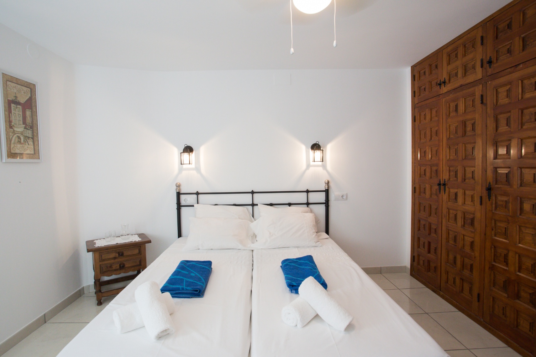 Dormitorio con camas individuales, cabecero de forja negra, apliques tipo farol y un amplio armario empotrado de madera con cuarterones tallados.