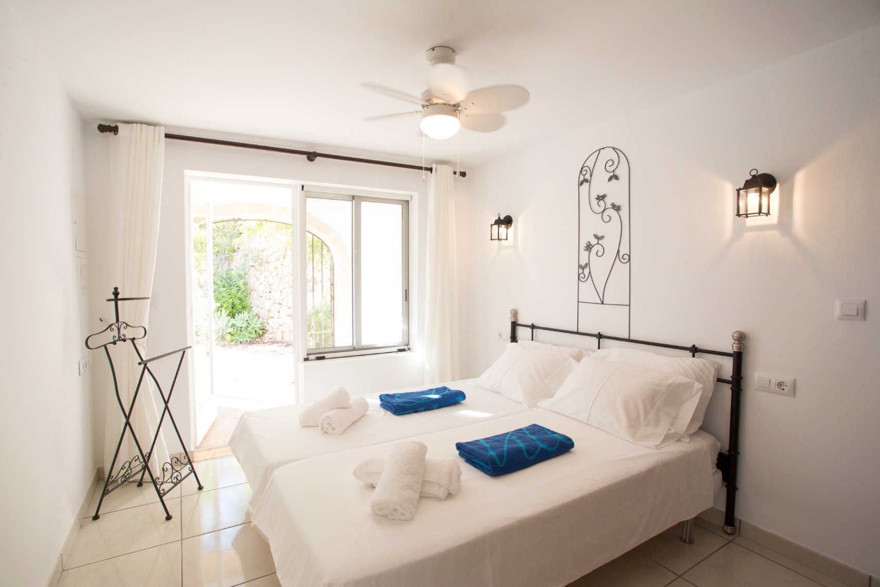 Dormitorio con dos camas individuales, cabecero de forja y apliques de pared. Dispone de ventilador de techo y acceso directo a un patio exterior con muros de piedra.
