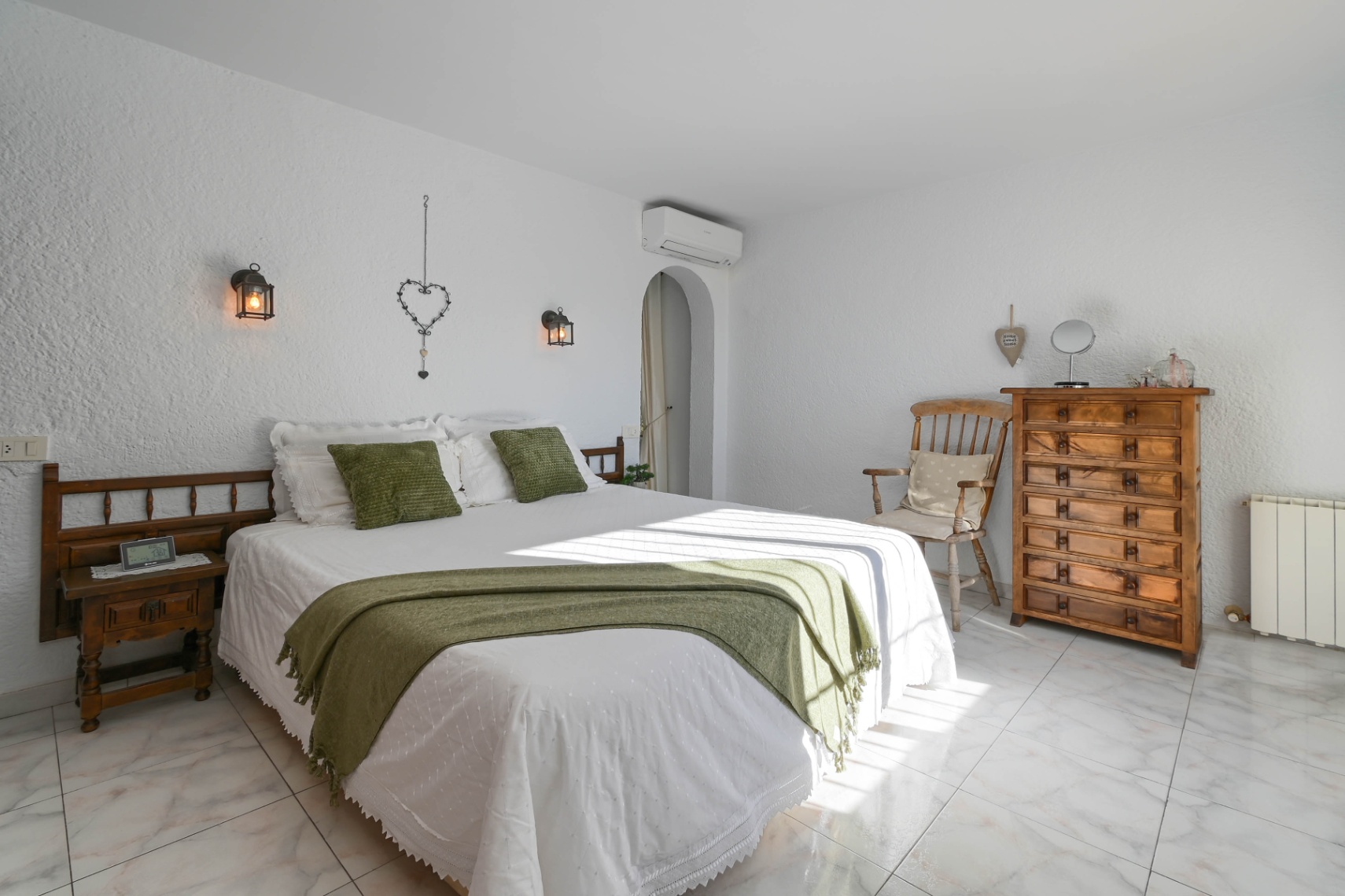 Dormitorio con paredes blancas texturizadas, suelo de mármol y muebles de madera rústica. Incluye iluminación de farol y textiles en tonos verdes.