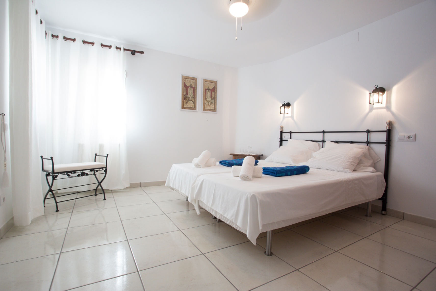 Dormitorio luminoso con camas individuales, cabecero de forja y suelos cerámicos. Incluye apliques de pared tipo farol y un banco de hierro junto al ventanal.