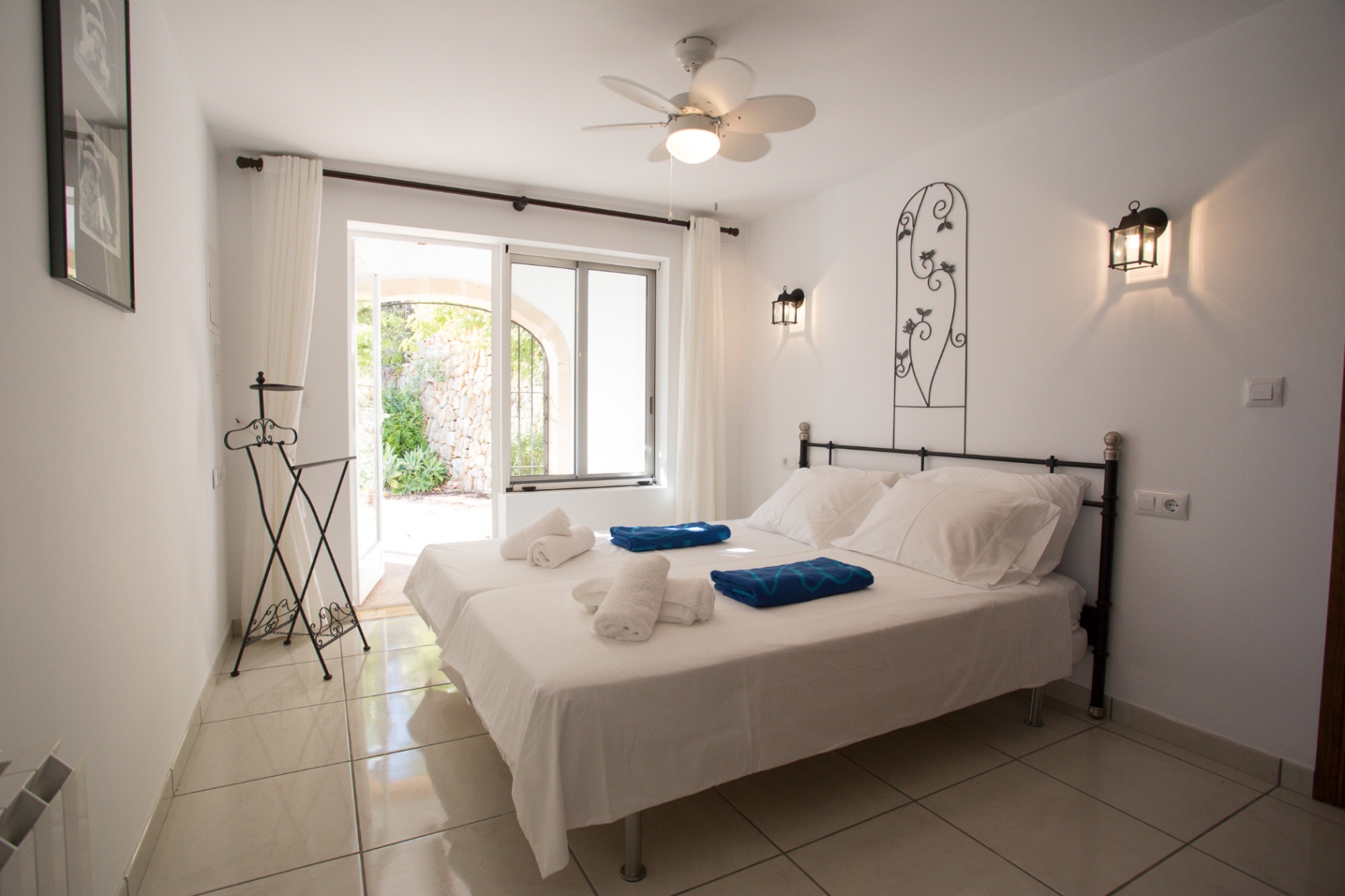 Dormitorio luminoso con camas individuales, detalles de forja y ventilador de techo. Una puerta acristalada conecta con un patio exterior de piedra natural.