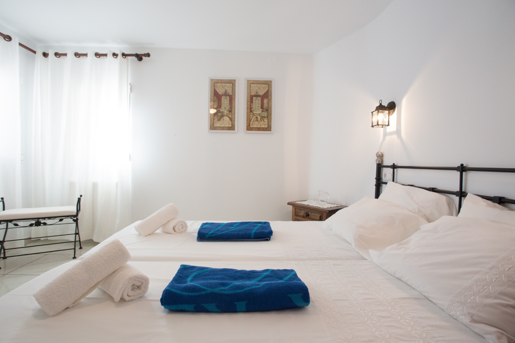 Dormitorio luminoso con camas individuales unidas, cabecero de forja negra y banco a juego. Incluye mesita de madera y luz natural a través de cortinas blancas.