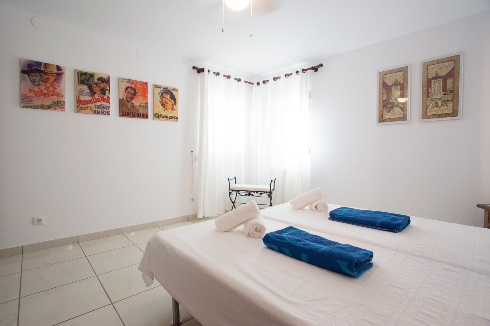 Dormitorio luminoso con dos camas individuales, carteles de cine clásico, suelos de baldosa y ventanas en esquina con cortinas blancas.