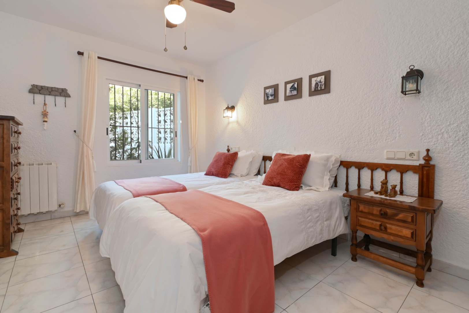 Dormitorio luminoso con dos camas individuales, detalles en color terracota y mobiliario de madera rústica. Incluye paredes de acabado rugoso y ventana con vistas al jardín.