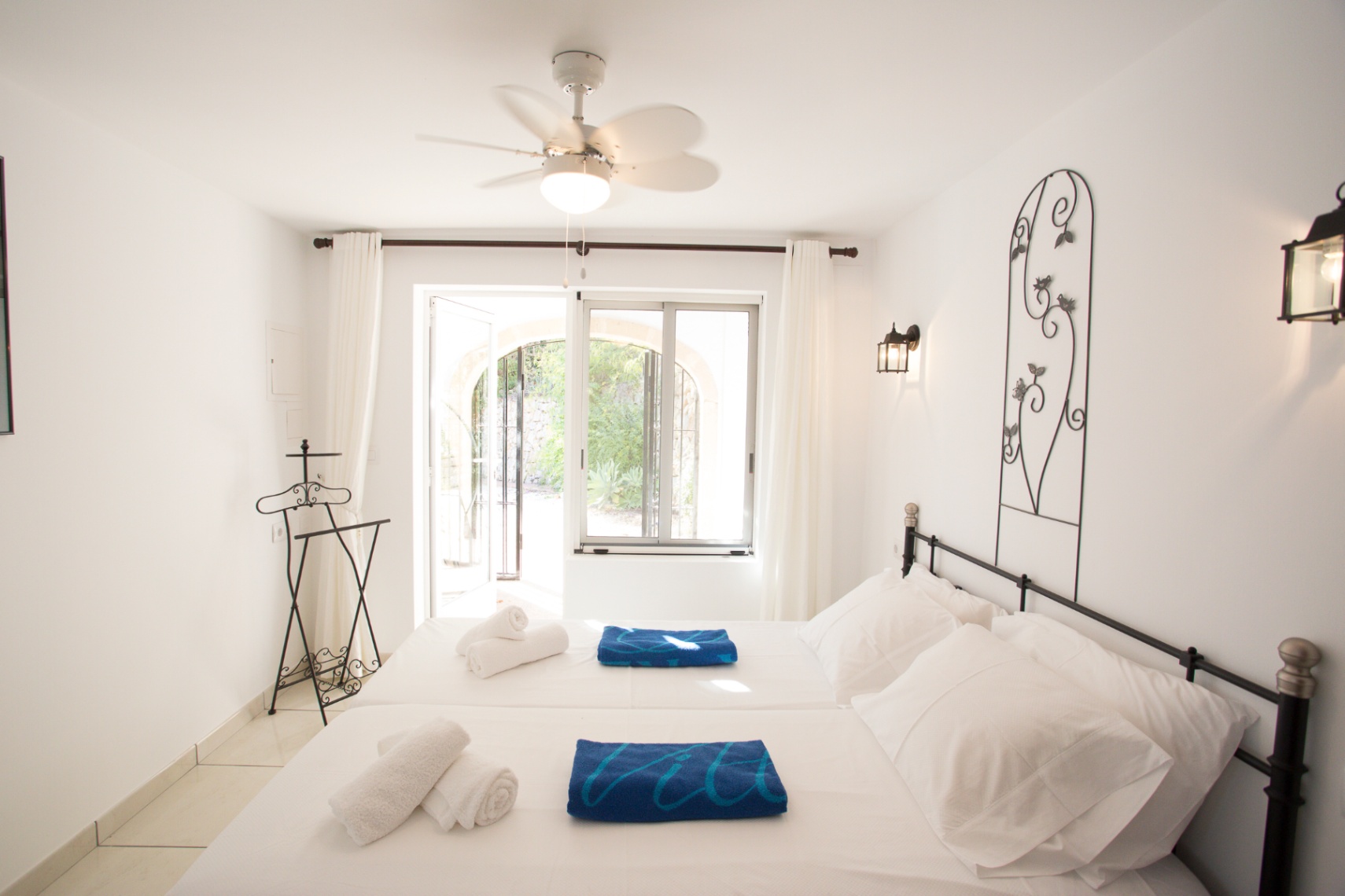 Dormitorio luminoso con paredes blancas, cabecero de forja y ventilador de techo. Dispone de acceso directo a un patio exterior a través de puertas acristaladas.