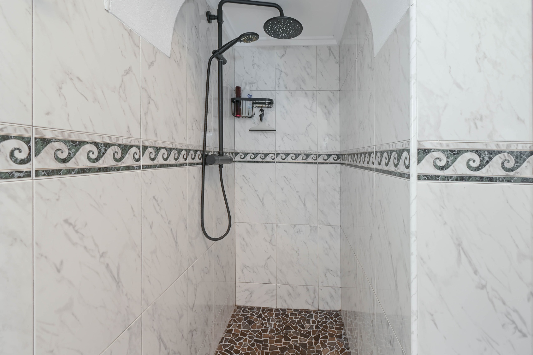Ducha de obra con azulejos efecto mármol, cenefa decorativa de ondas, suelo de mosaico de piedra y grifería negra mate con rociador de efecto lluvia.