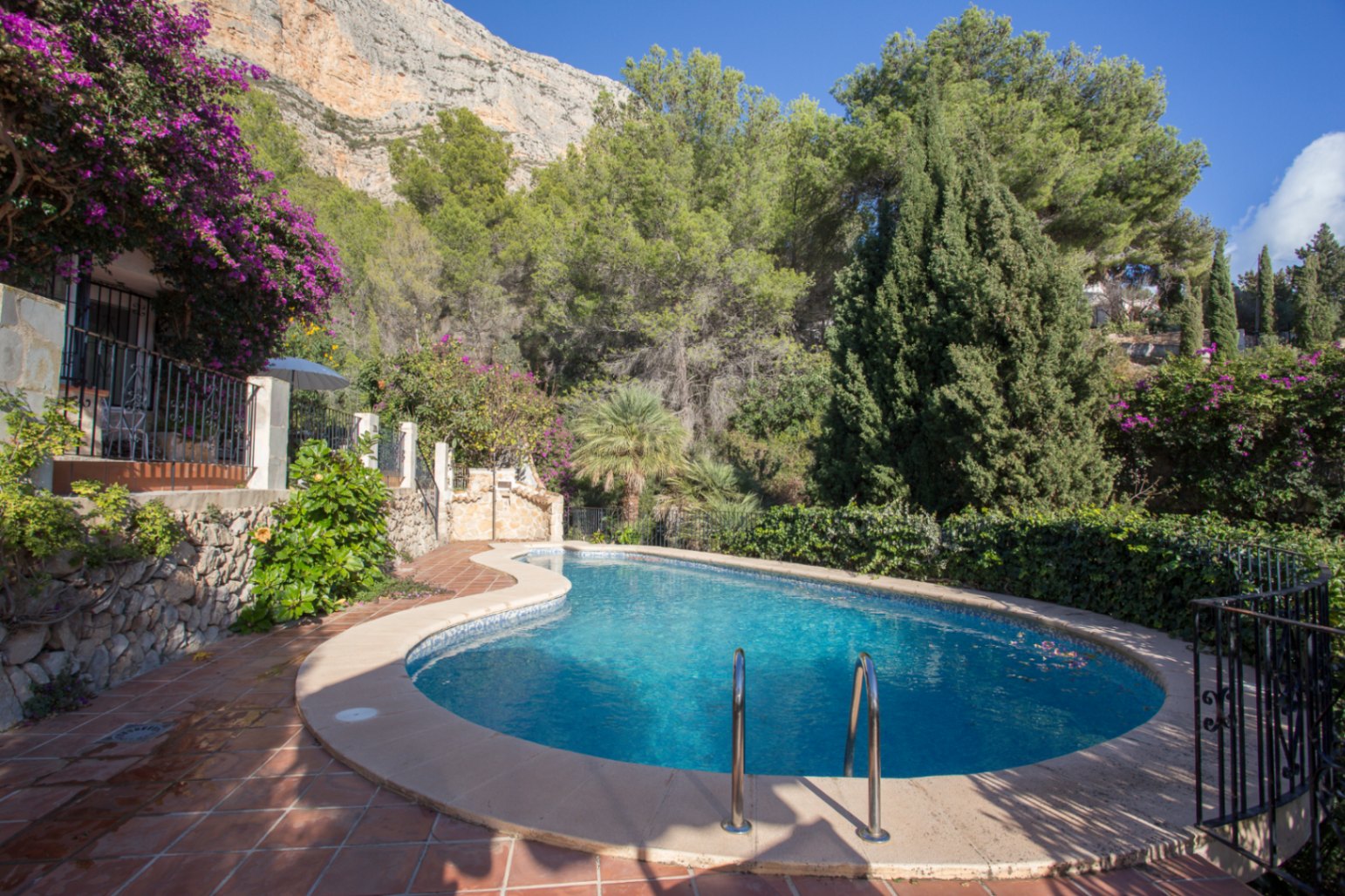 Piscina de forma curva con gresite azul, rodeada de suelos de terracota y vegetación mediterránea bajo una imponente pared de roca natural.
