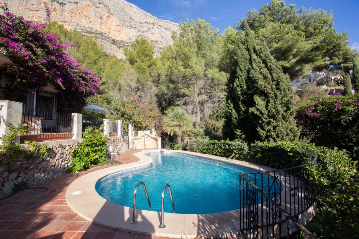 Piscina de forma irregular rodeada de una terraza de terracota y vegetación mediterránea, con vistas a un imponente acantilado de piedra caliza.
