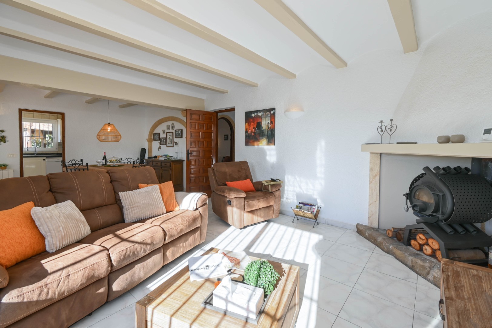 Salón con vigas de madera vistas, sofá marrón y una estufa de leña tradicional integrada en una chimenea. La estancia cuenta con suelos cerámicos y abundante luz natural.