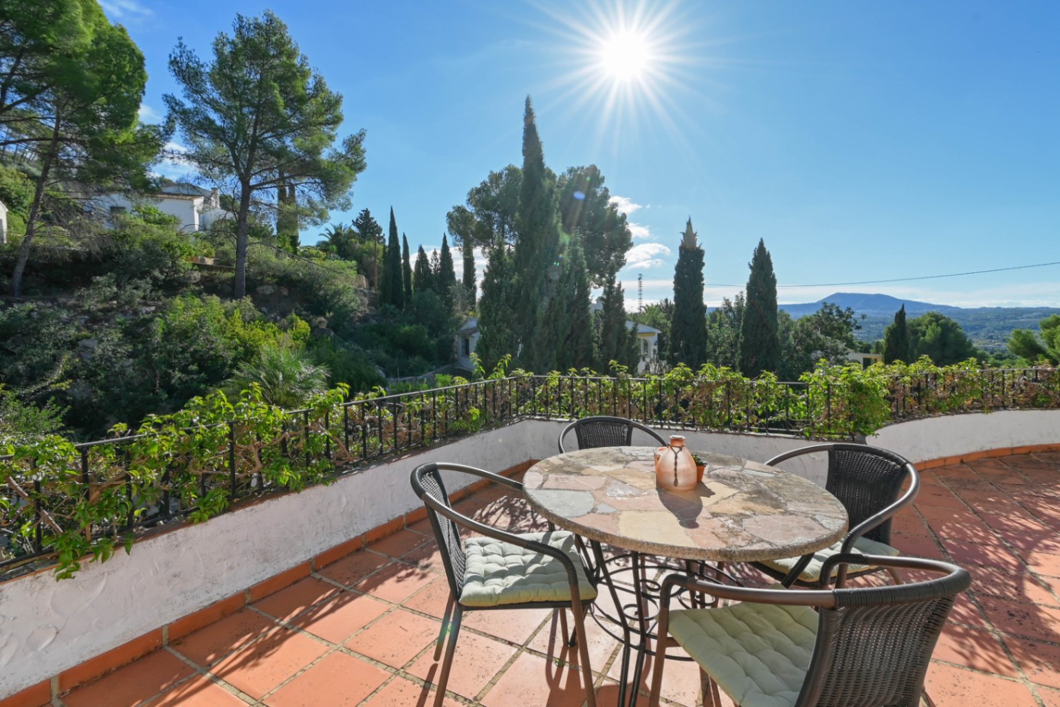 Terraza con baldosas de terracota, mesa de piedra y sillas de mimbre, con vistas a la vegetación mediterránea y montañas bajo un sol intenso.