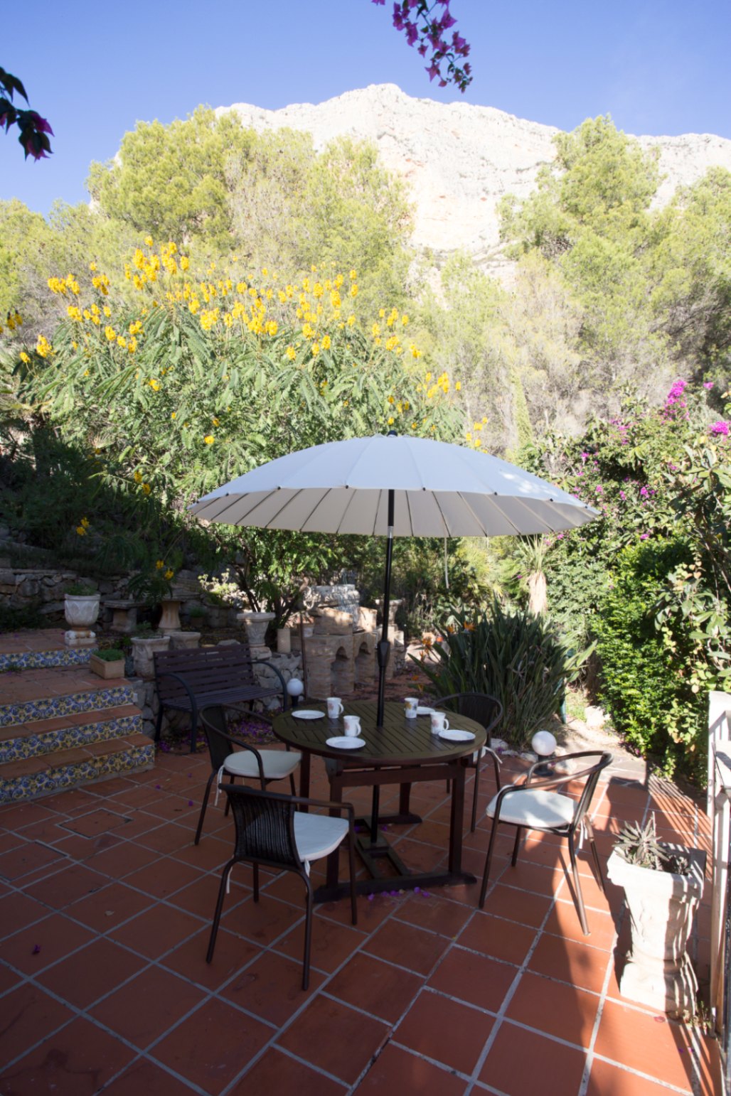 Terraza de baldosas de terracota con conjunto de comedor circular, sombrilla blanca y vistas a un jardín mediterráneo con fondo de montaña rocosa.