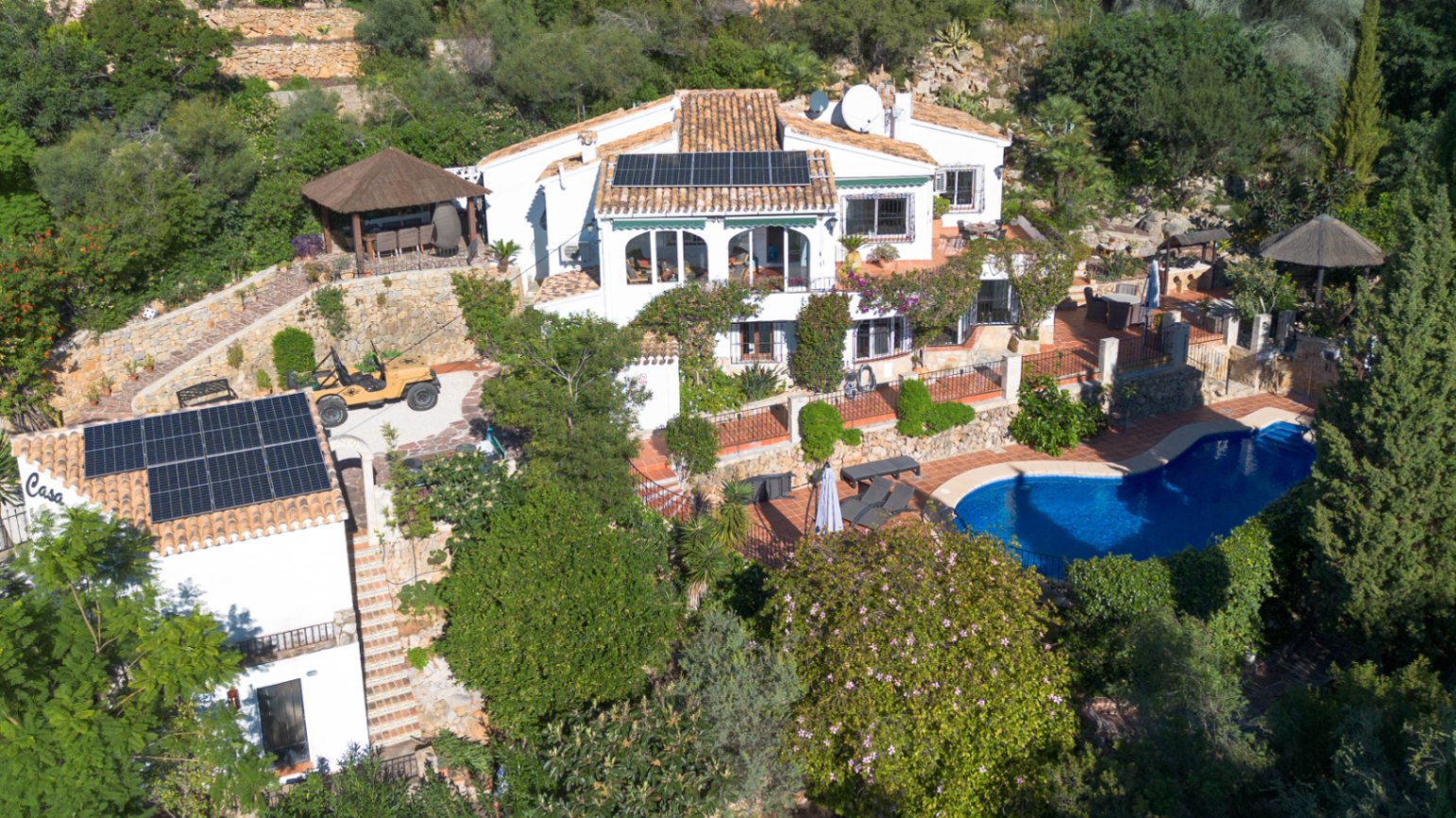 Vista aérea de una villa mediterránea con paneles solares, piscina de forma riñonada y jardines aterrazados con muros de piedra y vegetación exuberante.