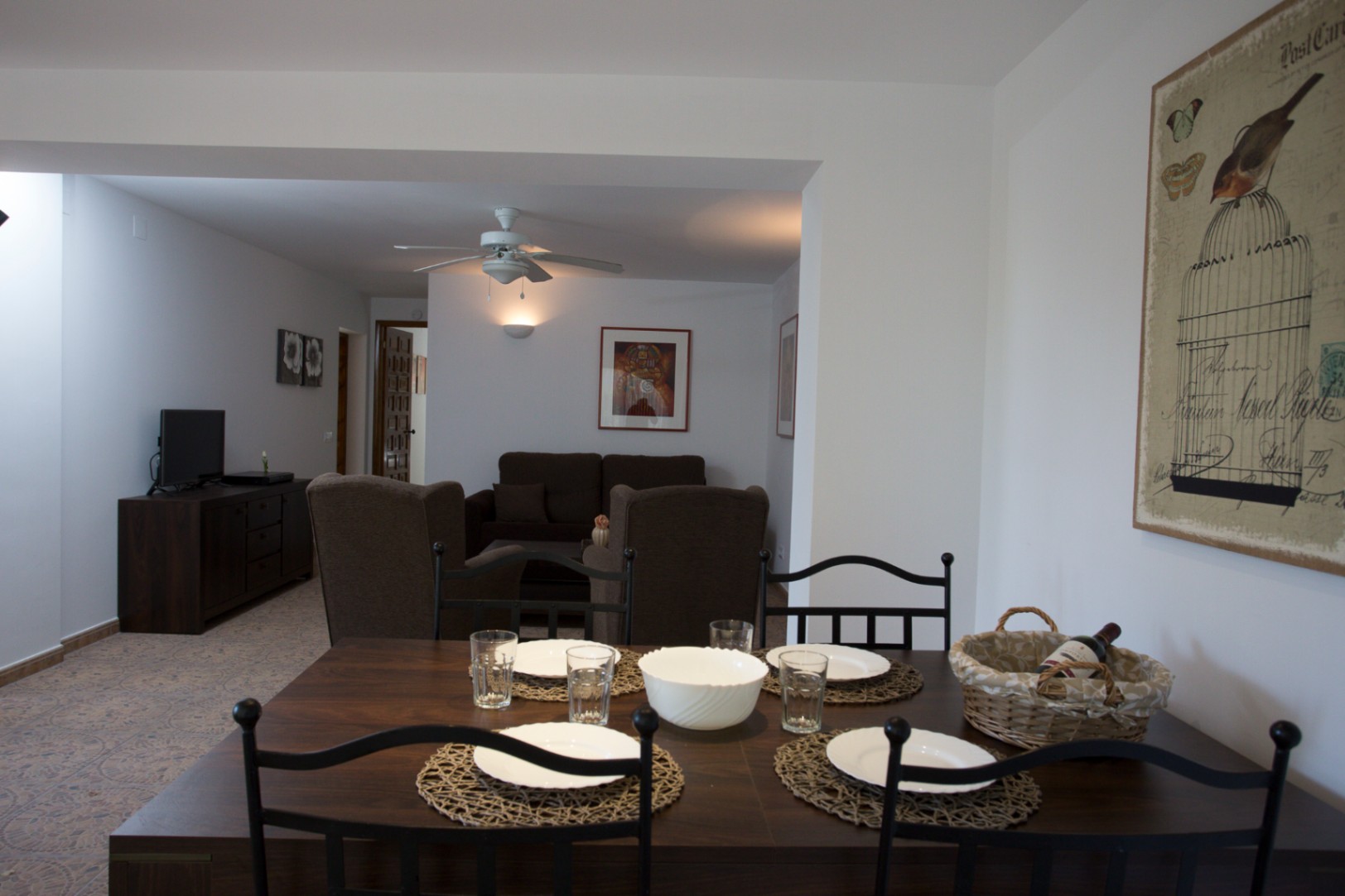 Zona de comedor y estar abierta con mesa de madera oscura, sillas de metal negro, suelos de baldosa con motivos y un salón con sofás marrones y ventilador de techo.