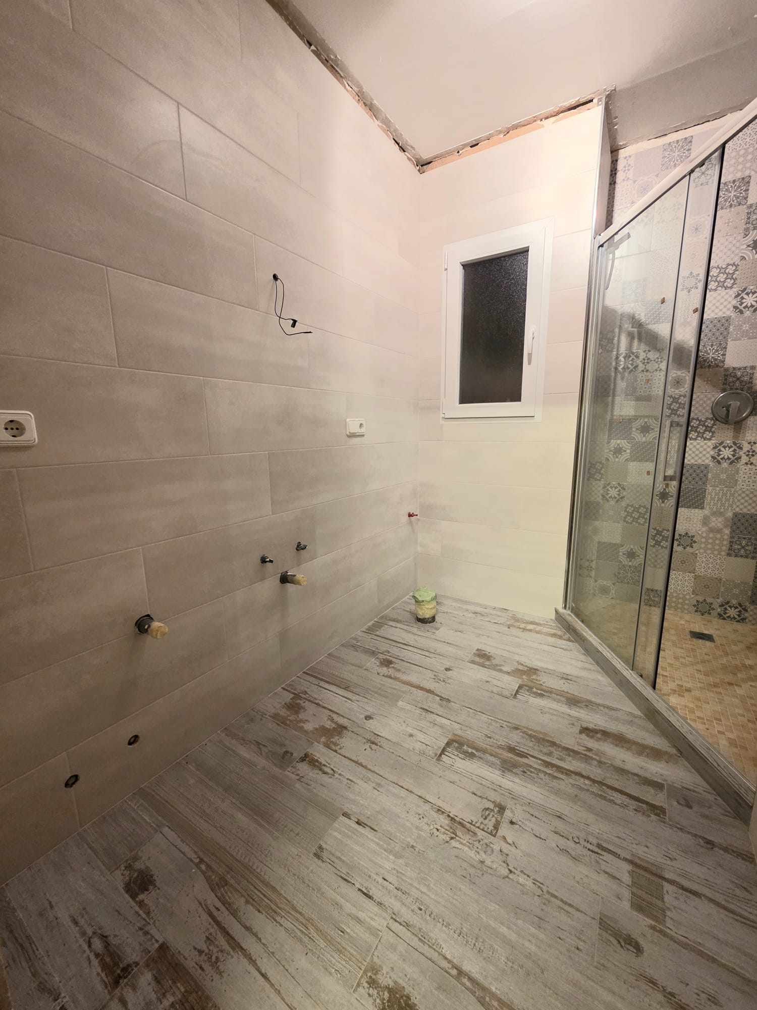 Baño en reforma con suelo cerámico imitación madera, paredes alicatadas en tonos neutros y zona de ducha con mampara de cristal y azulejos hidráulicos decorativos.