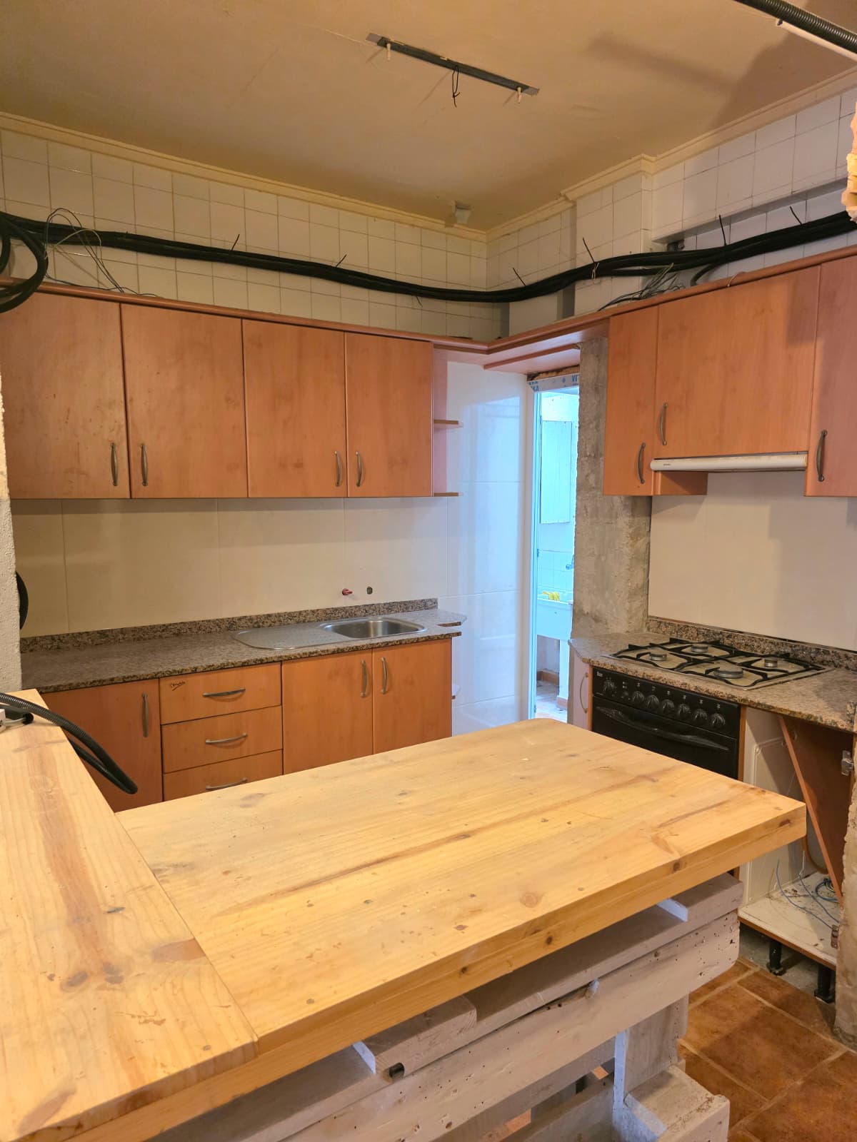 Cocina con armarios de acabado madera, encimeras de granito y una isla central de tablones. Incluye paredes alicatadas y cableado visto en la parte superior.